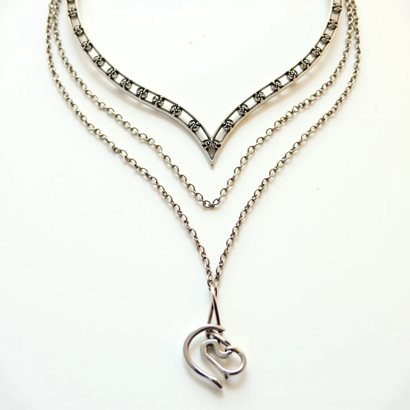 This layered necklace features a striking design composed of three distinct chains, each made from a shiny metal, likely silver or a silver-toned alloy. The topmost chain showcases a V-shaped centerpiece adorned with regularly spaced, small decorative elements that resemble inset stones, providing a textured appearance. The middle layer is a simple link chain that complements the complexity of the adjoining layers. The bottom chain elegantly suspends a pendant designed in an artistic form with intersecting loops, reminiscent of a stylized heart motif. The necklace is fastened with a classic lobster clasp, ensuring both style and functionality.