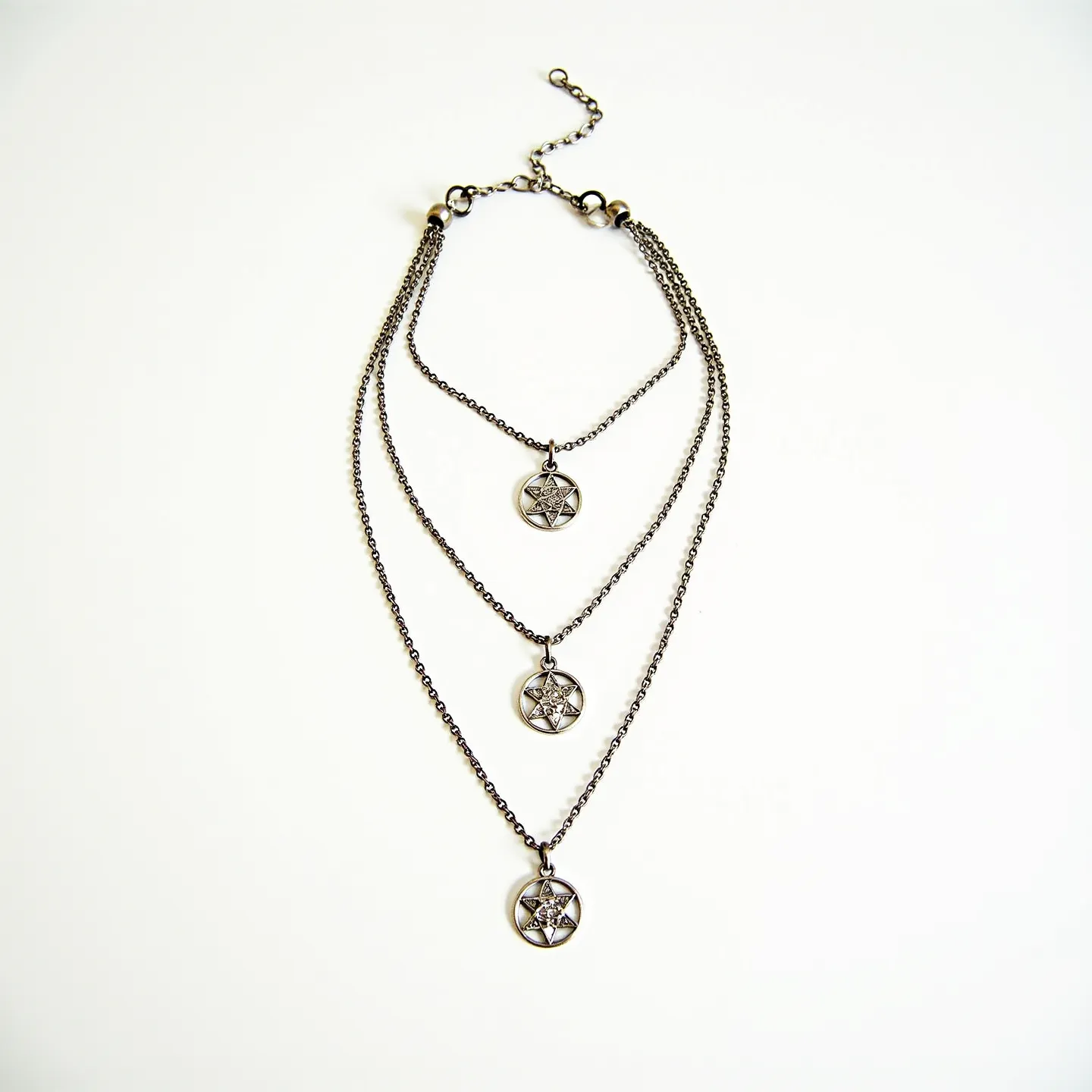 This layered necklace features a design with three chains of varying lengths, each adorned with a circular pendant showcasing a star motif. The chains appear to be crafted from a dark metal, possibly oxidized silver or a dark-toned alloy, imparting a vintage or gothic aesthetic. Each pendant is similarly composed, with raised star patterns that may be embellished with small, embedded stones for accentuation. The necklace is secured with an adjustable clasp, allowing for versatility in length, featuring a series of linked loops and a lobster clasp for easy attachment.