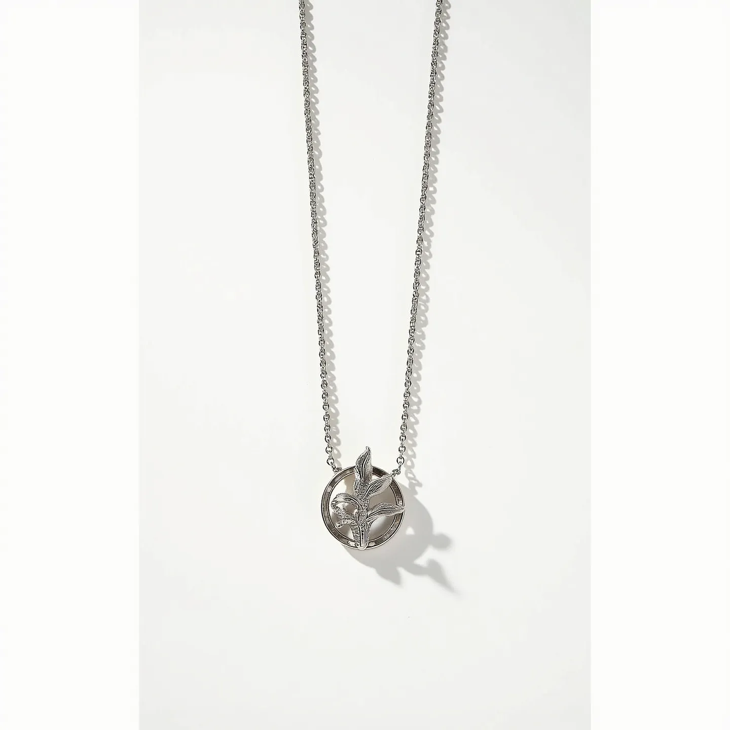 This layered necklace features a delicate chain made of a silver-toned metal, which lends it an elegant and subtle shine. At the center of the necklace hangs a distinctive pendant, designed with intricate cutout details that resemble botanical elements within a circular frame. The pendant itself is also made of the same silver-toned metal, enhancing the cohesive design. The necklace appears to be secured with a standard clasp, providing a simple yet secure attachment.