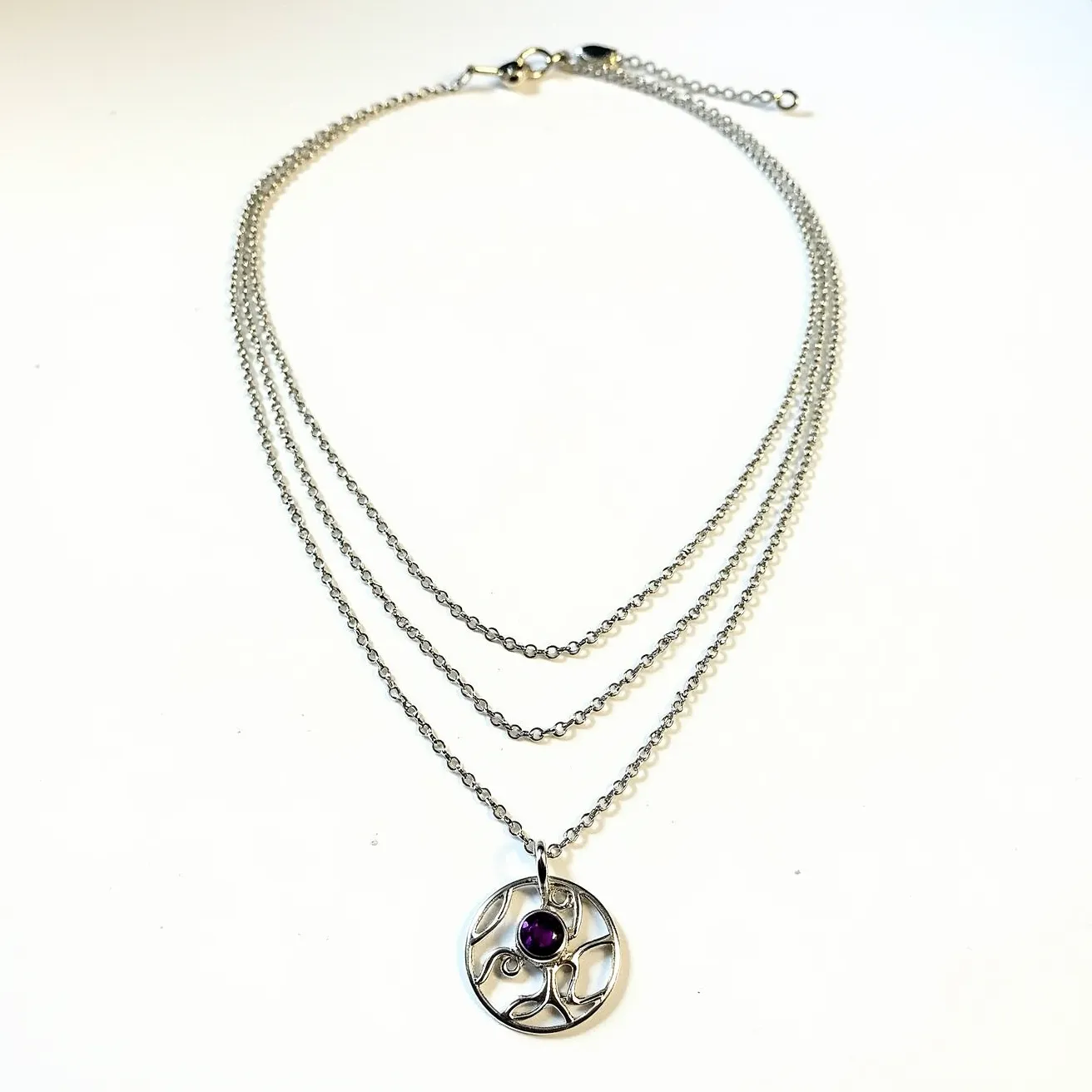 This layered necklace features multiple delicate chains, likely made from a silver-toned metal, giving it an elegant and subtle shine. The pendant is a circular openwork design, incorporating flowing, organic shapes with a central setting that holds a small, round-cut amethyst stone, offering a vibrant touch of purple. The amethyst is bezel-set, ensuring a smooth and polished appearance. The necklace is secured with a lobster clasp, which is attached to an adjustable chain section that provides flexibility in length.