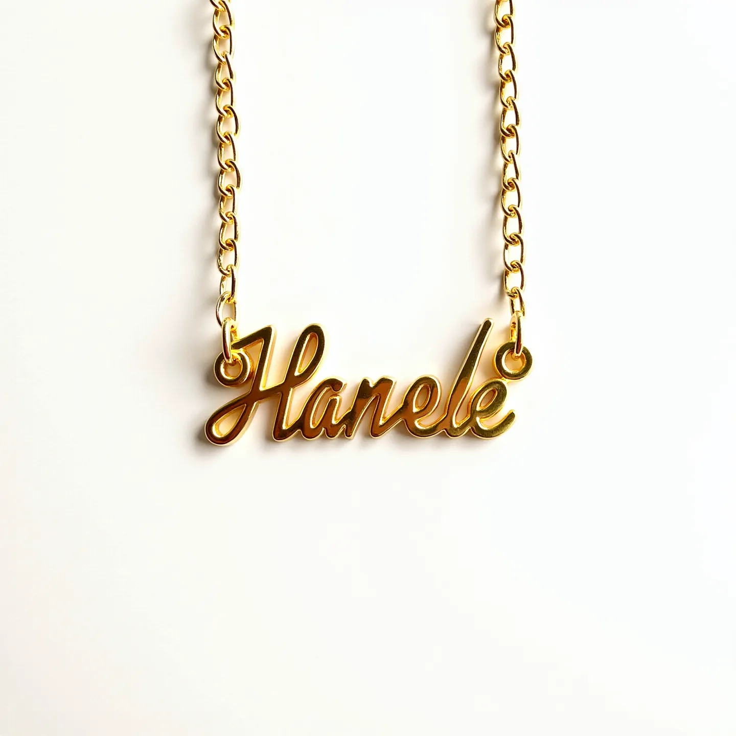 This letter necklace features the name "Hanele" crafted in a cursive script and is primarily composed of a shiny, gold-colored metal. The letters are smoothly interconnected, creating a continuous piece that elegantly displays the name. The nameplate attaches to a gold-toned chain with two loop connectors on either side, contributing to the necklace's cohesive aesthetic. The chain consists of evenly spaced, oval links that add a classic touch to the overall design.