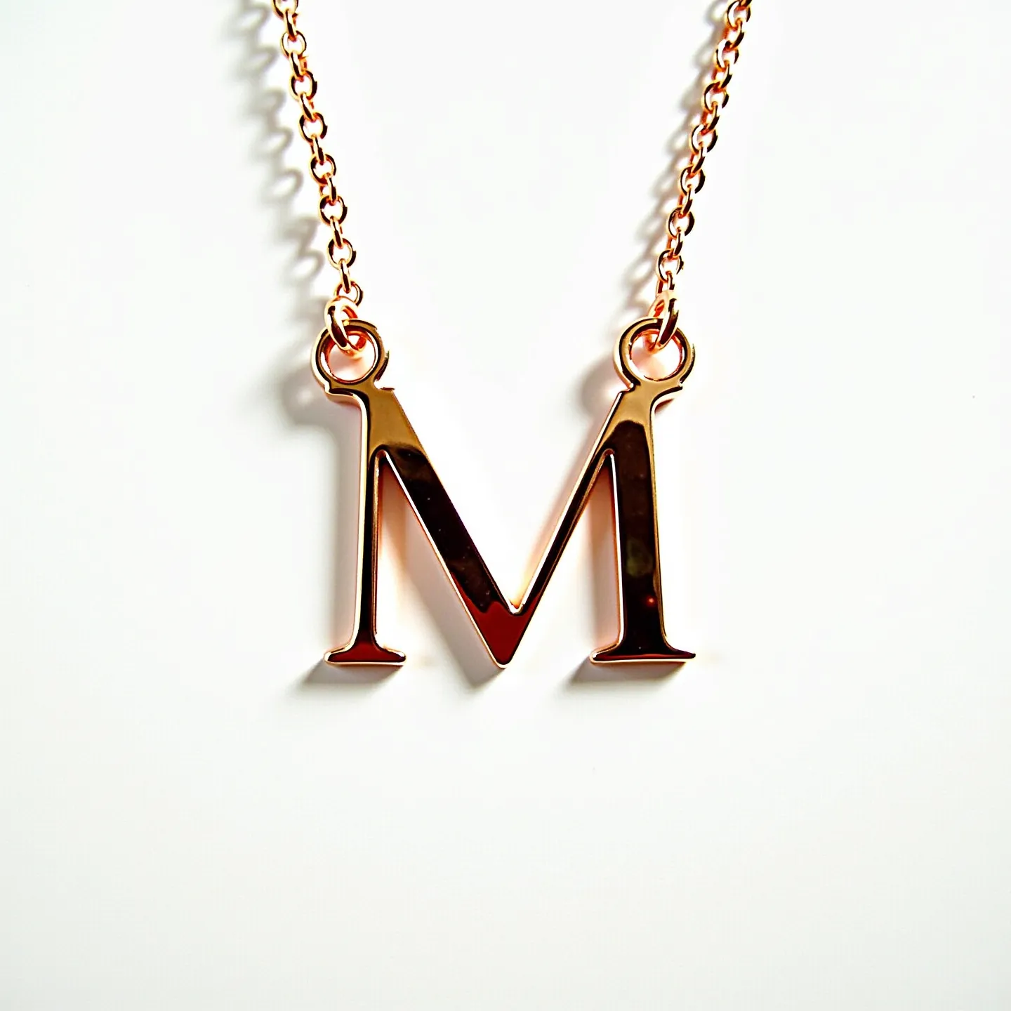 This letter necklace features a prominent "M" pendant crafted from a shiny, rose gold-toned metal, giving it a sleek and polished appearance. The chain appears to be composed of the same rose gold-toned material, matching the pendant for a cohesive look. The letter "M" is suspended by two loops at the top, securely attached to the chain. The chain is a standard link style, adding a classic touch to the design. The necklace is likely equipped with a typical clasp, such as a lobster or spring ring clasp, ensuring ease of wear and removal.