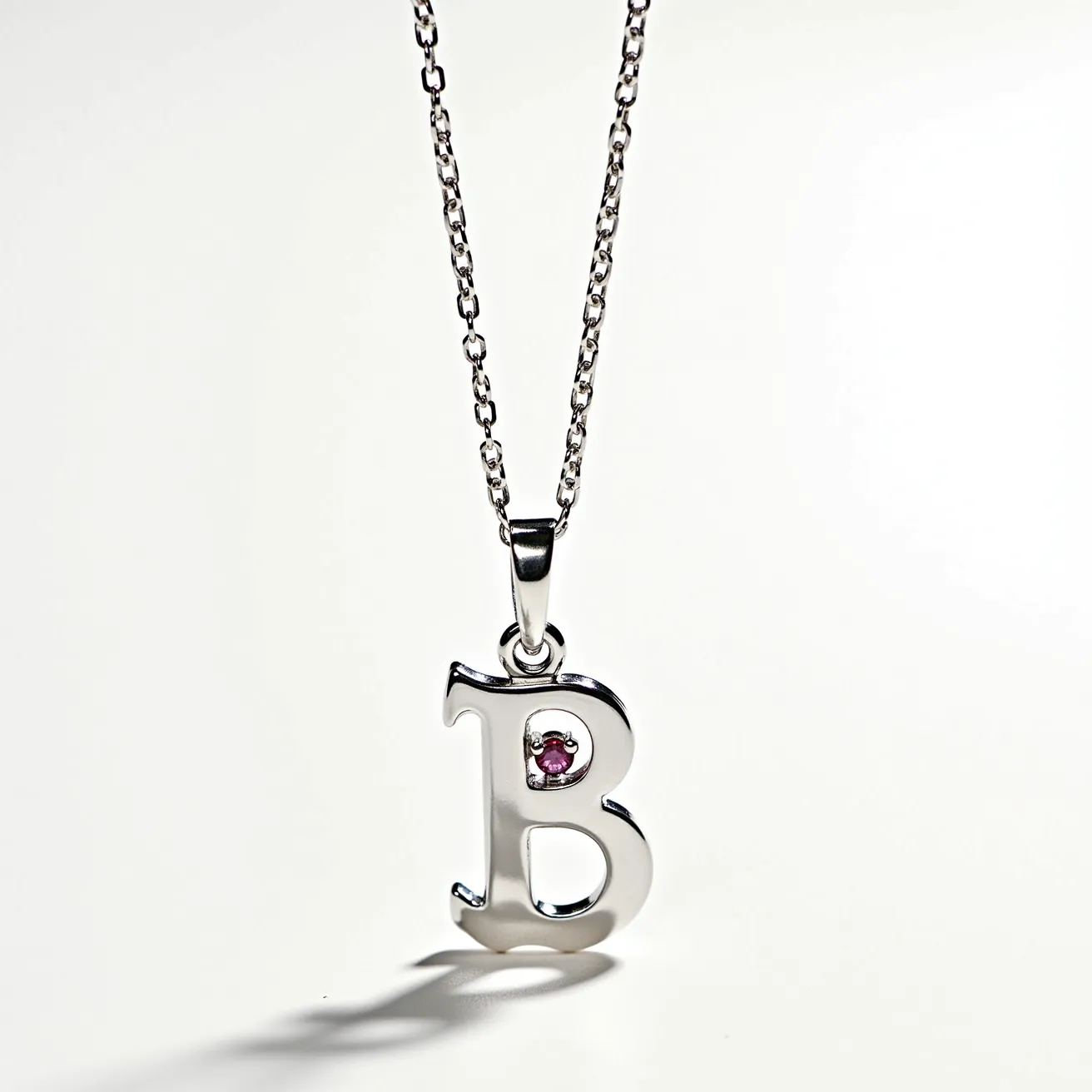 This letter necklace features a pendant shaped like the letter "B," crafted from polished metal, likely sterling silver or white gold. Embedded within the upper curve of the "B" is a small, round-cut pink gemstone, possibly a ruby or pink sapphire, set in a bezel setting that secures the gem with a smooth metal border. The necklace is suspended from a delicate chain with small, closely linked oval links that add to its refined appearance. The attachment passes through a simple yet elegant bail, which attaches the pendant securely to the chain, ensuring the necklace hangs gracefully.