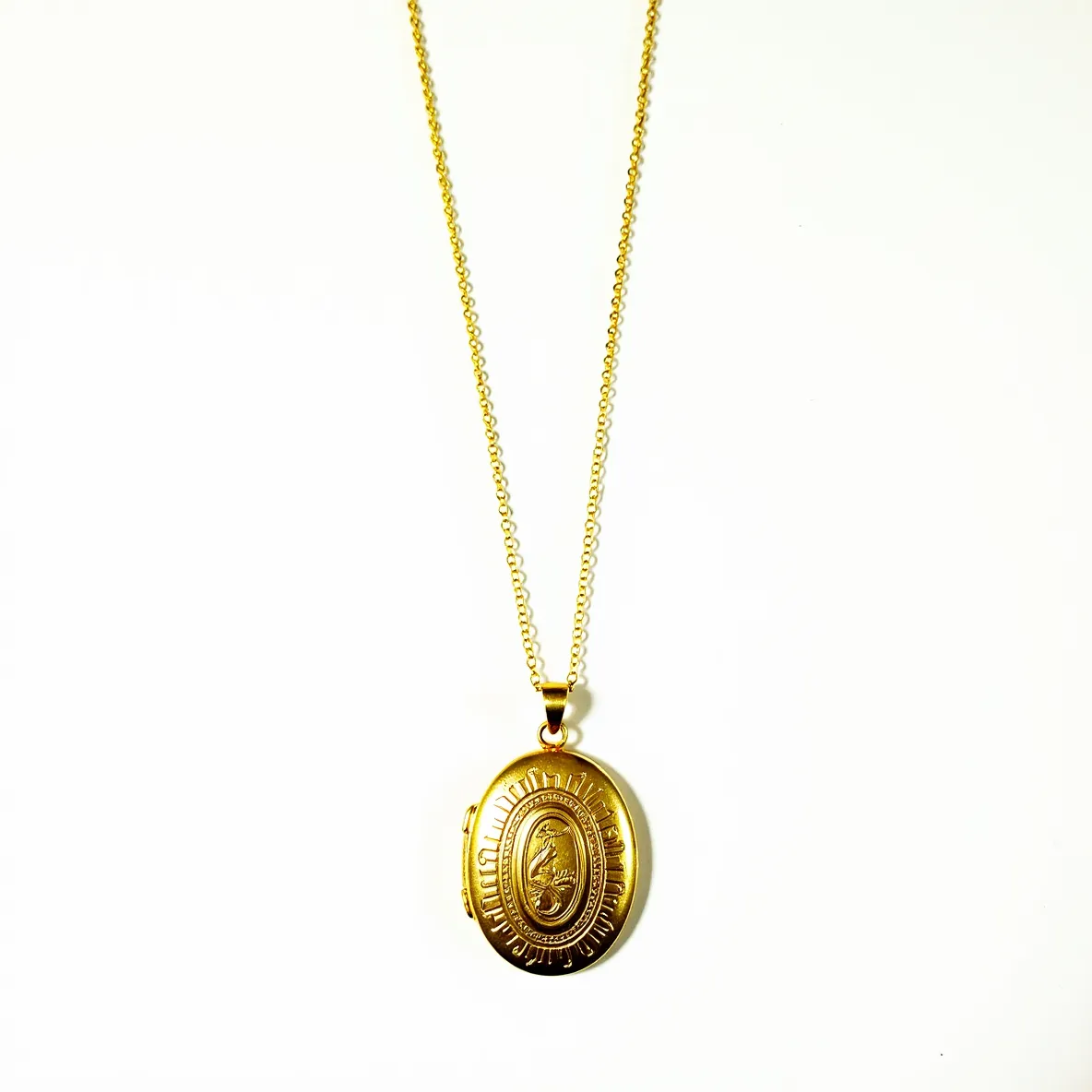 This locket necklace features an elegant oval-shaped locket crafted from gold-toned metal, showcasing intricate embossed detailing on its surface adding to its ornate design. The locket is attached to a delicate chain, which also appears to be made of matching gold-toned metal. The chain is composed of small, interconnected loops, contributing to its overall delicate appearance. The locket is suspended from the chain by a sturdy bail that allows it to hang smoothly. The necklace is likely secured with a standard lobster clasp, ensuring both functionality and security when worn. The locket’s distinctive design emphasizes a vintage aesthetic, blending both functionality and ornamental beauty.