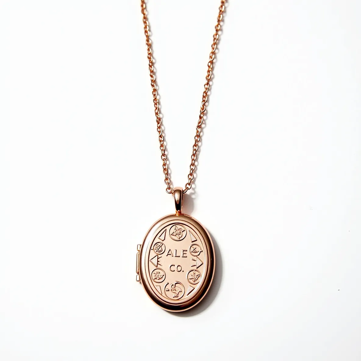 This locket necklace features an elegant oval-shaped pendant crafted from a rose gold-toned metal, giving it a warm and luxurious appearance. The front of the locket is engraved with intricate decorative symbols and the text "ALE CO," adding a personalized touch to the design. The pendant hangs from a finely linked chain, also in the same rose gold hue, which complements the locket beautifully. The locket includes a functional hinge on one side, allowing it to open and close securely. It is attached to the chain via a small loop at the top of the locket, ensuring it is held in place securely and gracefully.