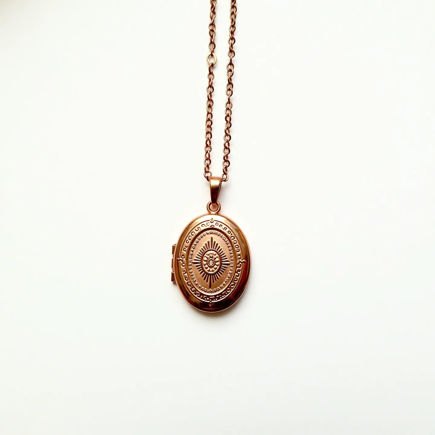 This locket necklace features a vintage design with an oval-shaped locket made of a warm, gold-toned metal. The front of the locket is intricately engraved with a sunburst pattern surrounded by a decorative border with scroll-like motifs. It hangs from a matching chain with oval links that complement the overall aesthetic. The locket is likely hinged for ease of opening. The necklace is attached with a simple loop bail, ensuring the locket hangs securely while adding to the classic style of the piece.