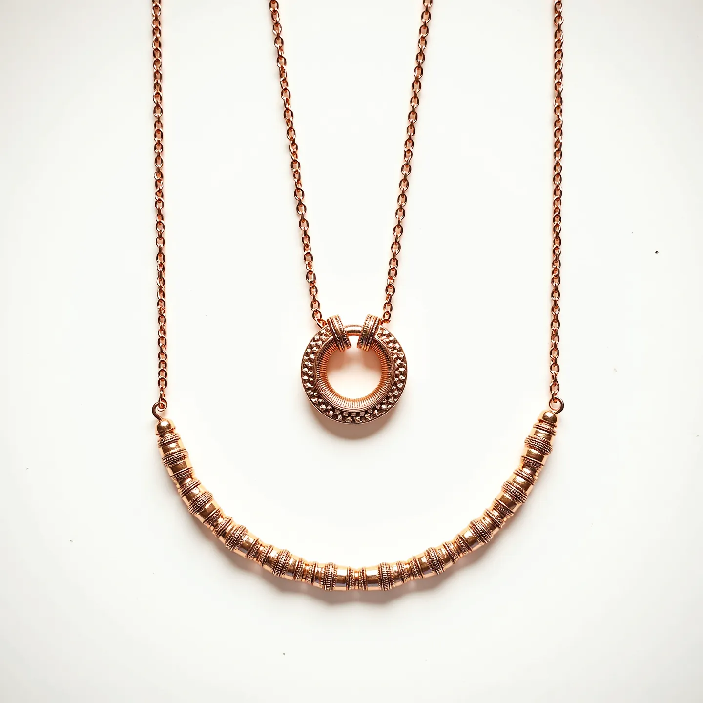 This long gold necklace features a delicate chain that supports two prominent pendant designs. The first pendant, located centrally, is a round, open circle adorned with intricate engravings and small bead-like details, adding a hint of sophistication. The second pendant is an elegantly curved bar composed of several cylindrical segments that are tightly arranged and detailed with engraved patterns, offering a textured appearance. The chain appears to be made from gold, giving the necklace a warm and luxurious look. The necklace is completed with simple loop clasps for easy attachment and wear.