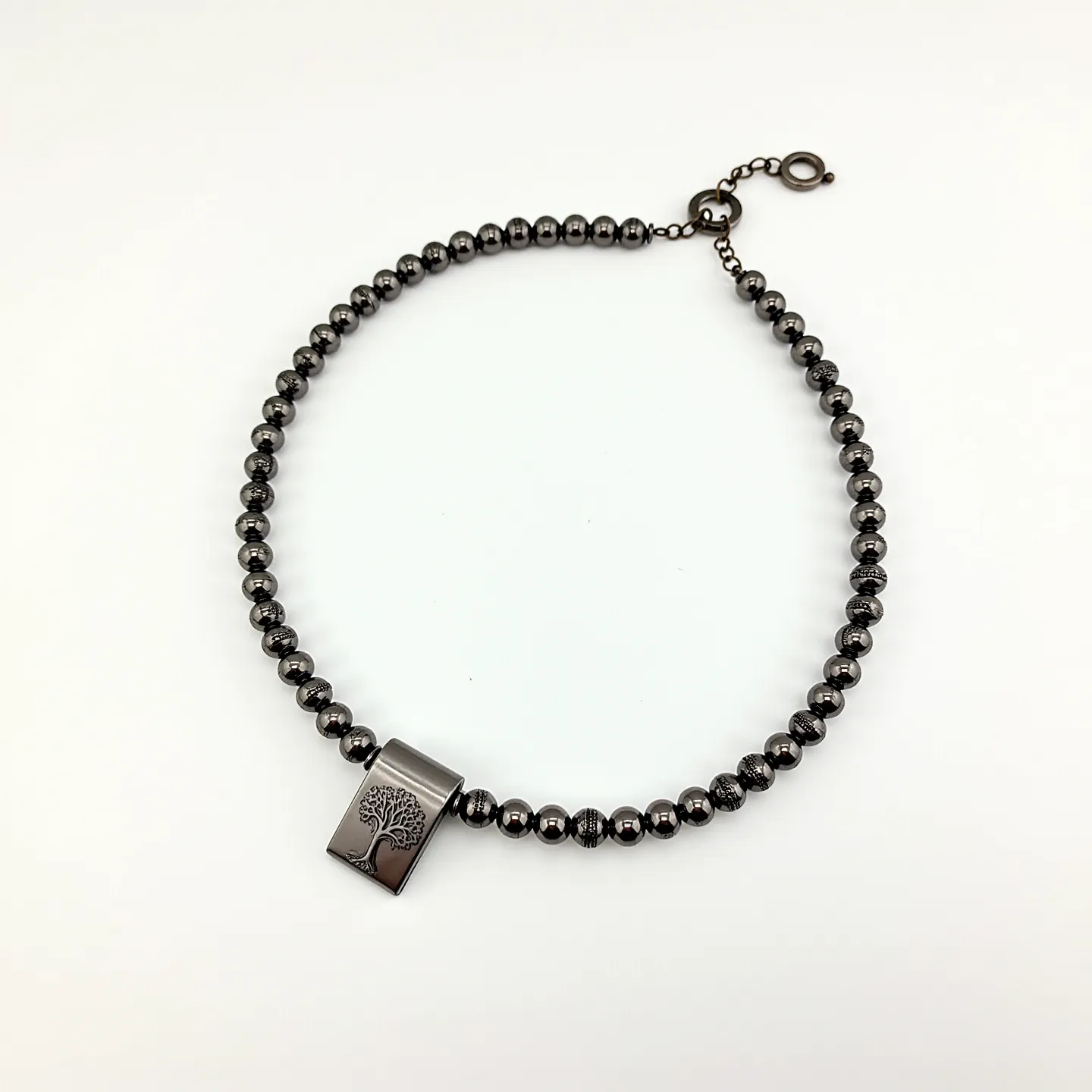 This men's beaded necklace features polished, spherical hematite beads, giving it a sleek, metallic sheen. A central rectangular pendant stands out, crafted from metal with a detailed etching of a tree, adding a distinct and meaningful design element. The necklace is secured with a metal clasp at one end, incorporating a hook-and-loop style for easy attachment and adjustment. The overall design combines functionality with a contemporary aesthetic, suitable for a variety of styles and occasions.