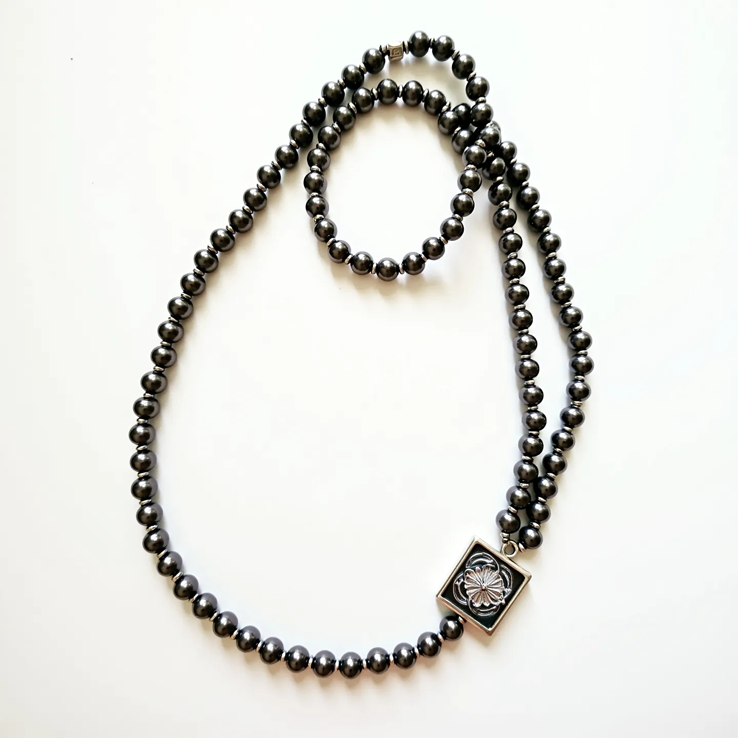 This men's beaded necklace features smooth, round hematite beads that are uniformly strung together, showcasing their metallic luster. At the center, a striking square pendant integrates a floral design in relief, adding a touch of elegance. The pendant appears to be set in a silver-tone metal, complementing the dark hue of the beads. Completing the necklace is a small, discreet metallic clasp, ensuring a secure and seamless closure.