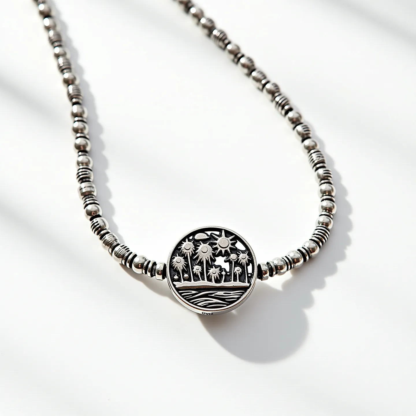 This men's beaded necklace features a combination of polished silver beads with varying sizes and designs, including spherical and cylindrical shapes. The central pendant is a round medallion with an intricate engraving of a tropical scene that includes palm trees and a setting sun, adding an artistic focal point. The beads and pendant are likely made of sterling silver, giving the necklace a classic metallic sheen. The necklace is designed to be worn easily with a discreet clasp at the back, maintaining the overall sleek and stylish aesthetic.