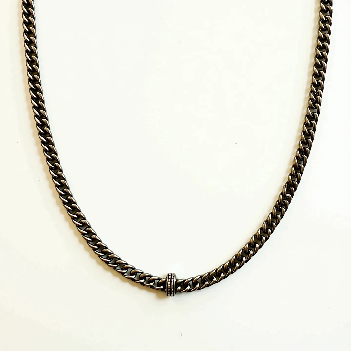 This men's necklace features a sleek, polished metal chain with a dark, possibly oxidized, finish that gives it a modern and stylish appearance. The chain links are tightly woven, suggesting a durable and substantial construction. At the center, there is a small, cylindrical bead with fine engravings, adding an element of intricate detail to the overall design. The necklace appears to be unadorned with gemstones, focusing instead on a minimalist aesthetic. The clasp or method of attachment is not visible, but the uniform design of the necklace suggests a seamless connection.