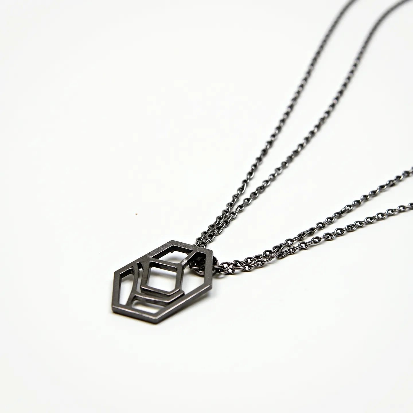 This men's necklace features a double-chain design made from a dark metal, possibly stainless steel or a similar alloy, giving it a modern and masculine look. At the center is a geometric pendant with a hollow 3D hexagonal shape, accentuating contemporary style. The chains are finely linked, adding an elegant touch, while the pendant's design adds a unique visual interest. Though the clasp isn't visible, the overall design suggests a piece crafted for both durability and style, ideal for a sleek, minimalist aesthetic.