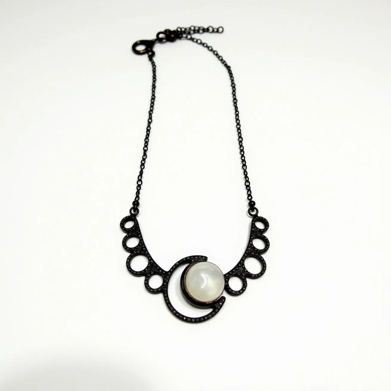 This celestial moon necklace features a striking design with a dark metal chain and an intricate pendant. The centerpiece showcases a polished, round moonstone, set in a pronged bezel that complements its glossy surface. The moonstone is framed by a black crescent, which is adorned with a series of small circular accents that add a textured dimension to the piece. The necklace is secured with a lobster clasp, allowing for adjustable length and a comfortable fit.