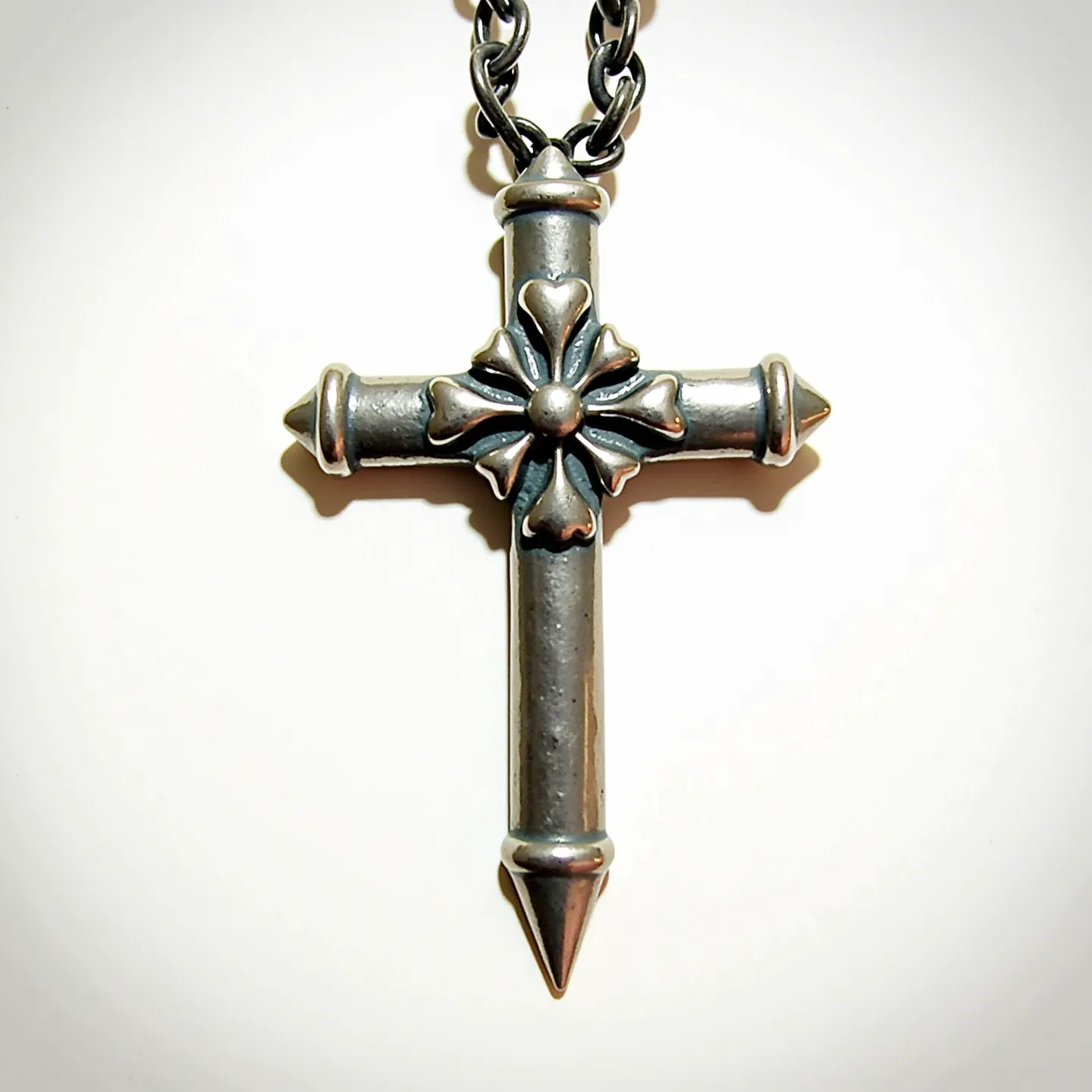 This nail cross necklace features a robust design with a metallic finish, likely crafted from stainless steel or a similar alloy, emphasizing durability and a rugged aesthetic. The cross is designed to resemble nails, with pointed ends and a symmetrical form. At the intersection of the cross, there is a decorative element resembling a flower or ornate motif, adding a touch of intricacy to the otherwise minimalist design. The pendant is suspended from a sturdy chain, connected through a simple loop at the top of the cross. The chain itself appears to be of the same metallic material, offering a cohesive and continuous style.