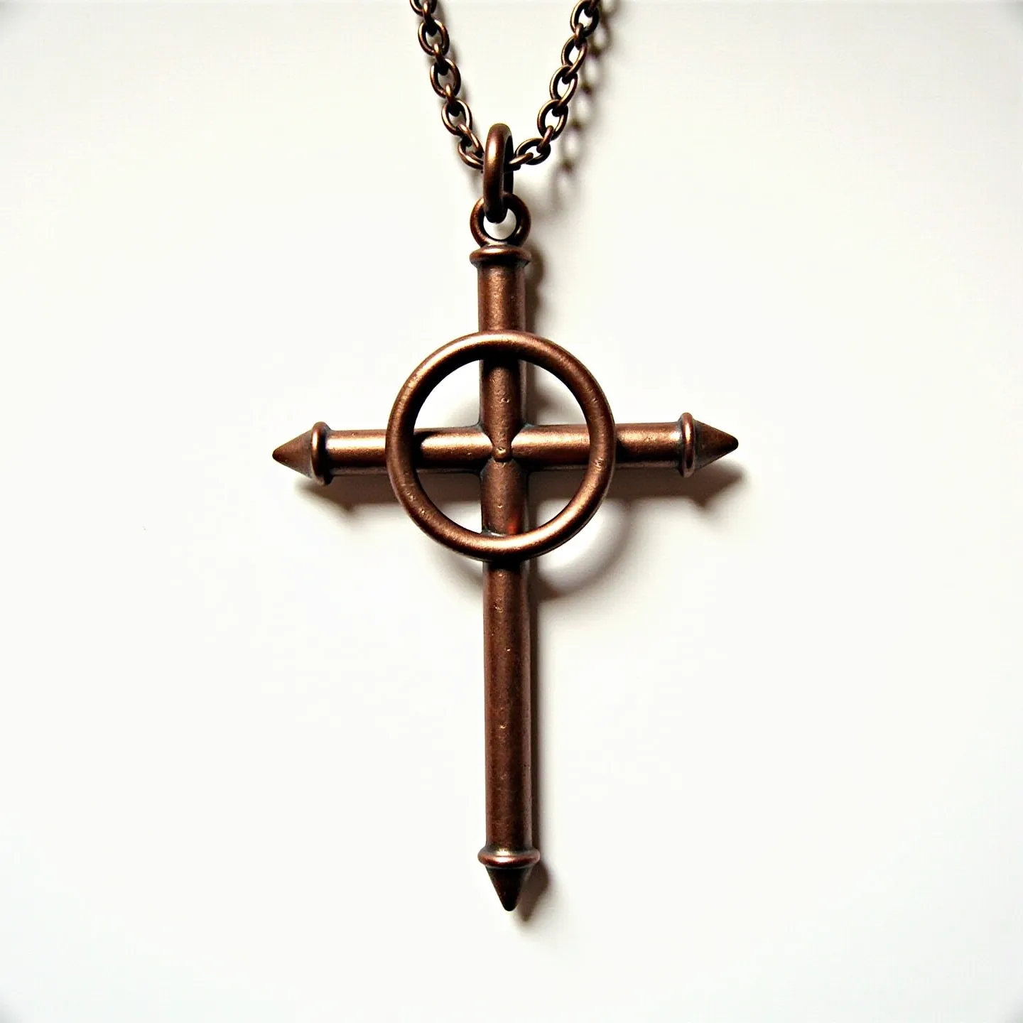 This nail cross necklace features a rustic design made of metal that resembles iron or antiqued brass, giving it a robust and vintage appearance. The cross is formed by two elongated nails intersecting, with the heads and points prominently visible, adding to its rugged aesthetic. At the intersection, a circular band encircles the center, enhancing the design with a touch of complexity. The cross pendant is attached to a sturdy chain via a simple ring loop, providing secure connectivity. The lack of gemstones or intricate settings contributes to the necklace's straightforward yet striking design.