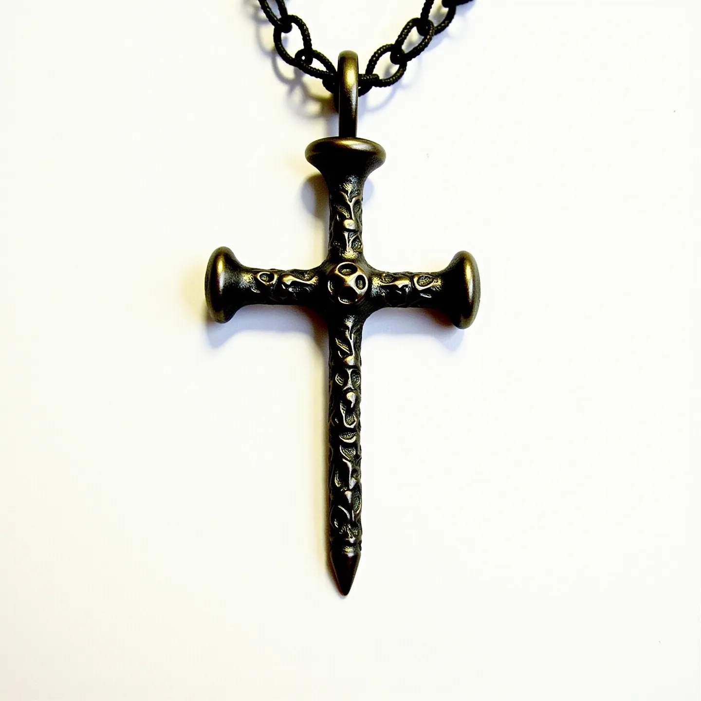 This nail cross necklace features a pendant shaped like a cross, crafted to resemble nails, with an antique brass or bronze finish. The design includes intricate detailing on the surface, reminiscent of decorative carvings or engravings. The cross is suspended from a cord that appears to be made of a braided or twisted black material, possibly leather or fabric. The attachment involves a loop at the top of the pendant, through which the cord is threaded, providing a secure connection. The necklace's design combines a rugged aesthetic with symbolic elements, creating a distinctive piece of jewelry.