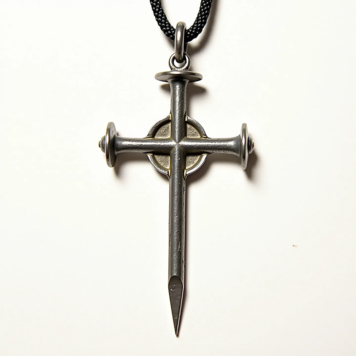 This nail cross necklace features a striking design composed of metallic nails forming the shape of a cross, with two nails crossing over one vertical nail. The nails appear to be made of a silver-toned metal, giving the piece a rustic and industrial aesthetic. Each nail is detailed with realistic features, such as the rounded heads and pointed ends. A circular element is situated at the intersection, providing an additional layer of visual interest. The pendant hangs from a black cord with a robust, woven texture, likely attached by a metal loop that secures the cross to the necklace. The combination of materials and design elements contributes to a bold yet understated appearance.