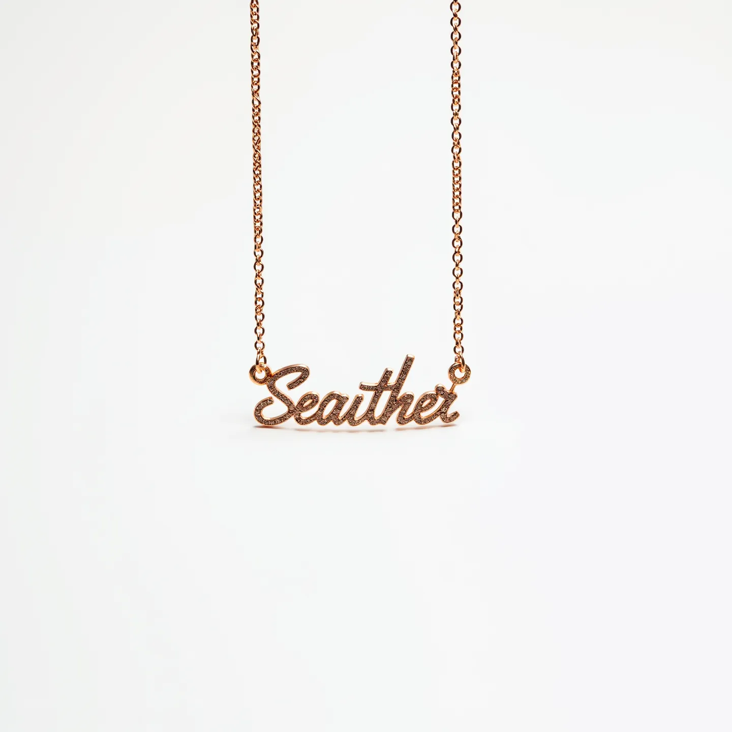 This name chain necklace features the name "Seanther" elegantly crafted in a script style, with each letter smoothly connected. Made from a warm-toned metal, likely gold or a gold-colored alloy, the piece provides a luxurious appearance. The chain is a delicate link style, which complements the intricacy of the nameplate. The letters are adorned with small, sparkling gems that resemble diamonds, cut in a way that catches light beautifully. The gems are meticulously set within the letters, enhancing their legibility and adding a touch of glamour. The necklace fastens with a standard lobster clasp, ensuring both security and ease of wear.