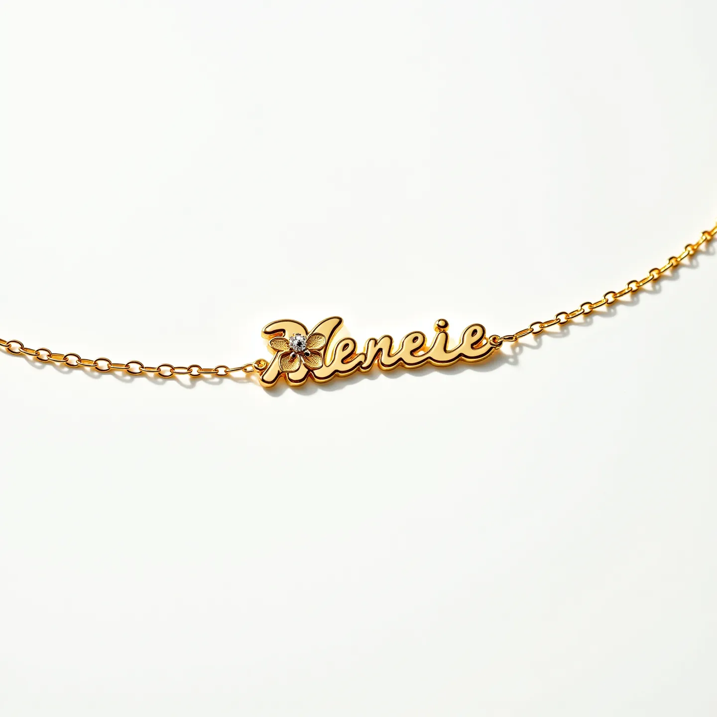 This name chain necklace is crafted from a gold-tone metal, featuring a delicate chain that elegantly holds the personalized nameplate. The nameplate showcases a script-style font adorned with a small, flower-like embellishment. At the center of the flower, there is a single, round-cut gem set in a simple, prong setting, adding a subtle sparkle to the design. The chain links are uniformly small and finely interlocked, offering both flexibility and strength. The necklace likely utilizes a standard lobster clasp for easy fastening and a secure fit around the neck.