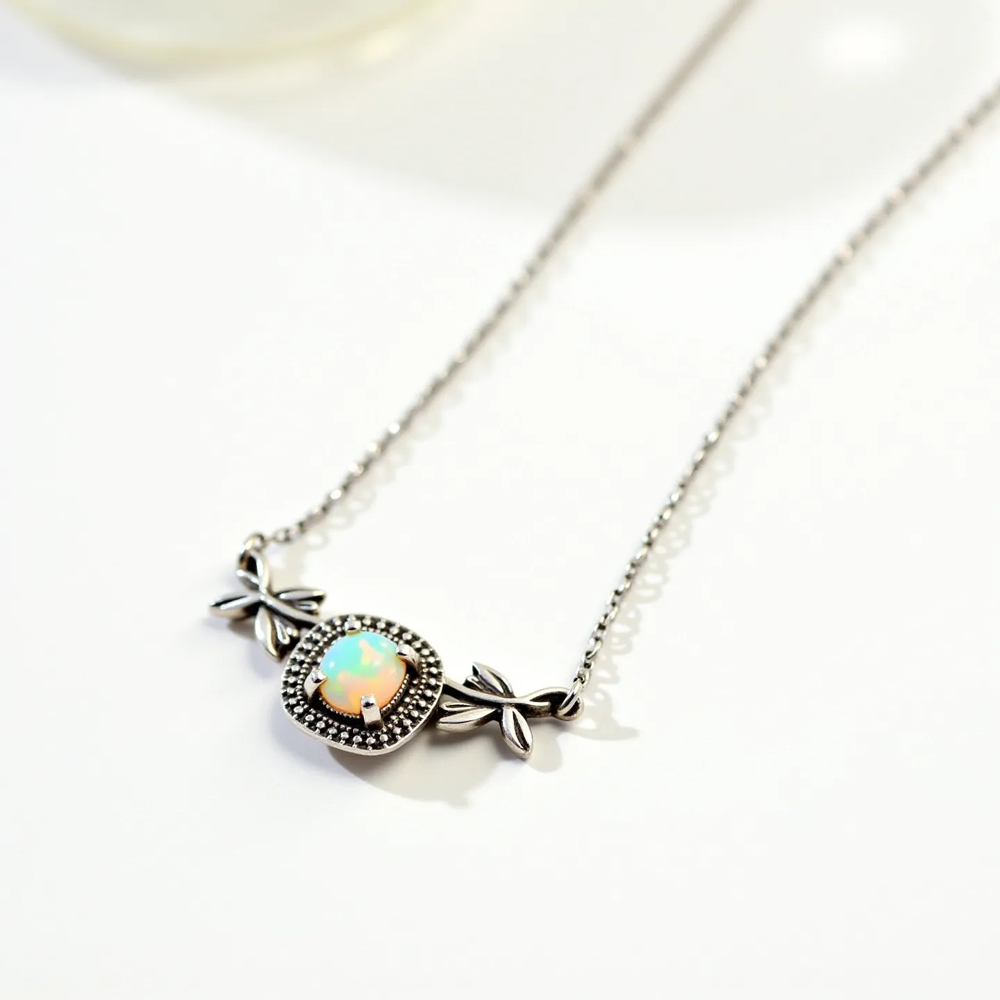 This opal necklace features a central opal gemstone with a captivating play of colors, cut in a cabochon style to highlight its iridescent qualities. The opal is set in a decorative bezel setting adorned with a detailed border that enhances its beauty. The necklace is crafted with a delicate metal chain, likely silver, contributing to its elegance. Flanking the opal are ornamental elements resembling bows or leaves, adding a touch of sophistication to the design. The necklace is completed with a clasp that ensures secure attachment around the neck, making it both a beautiful and functional piece of jewelry.