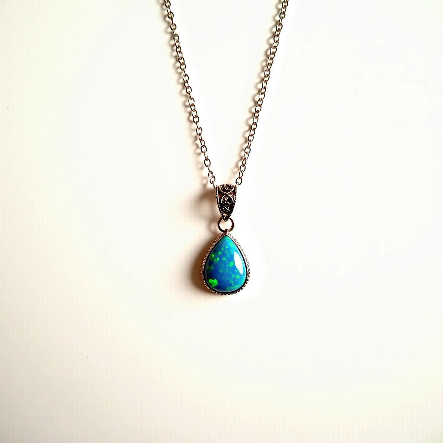 This opal necklace features a teardrop-shaped opal gemstone set in a simple bezel setting that highlights the opal's vibrant play of colors, predominantly blue with flashes of green. The opal is attached to a delicately designed metal bail that includes detailed ornamental patterns, adding an extra touch of elegance. The chain appears to be made of a fine metal, likely silver or silver-colored, complementing the opal's cool tones. A standard lobster clasp secures the necklace, providing both functionality and a seamless look.