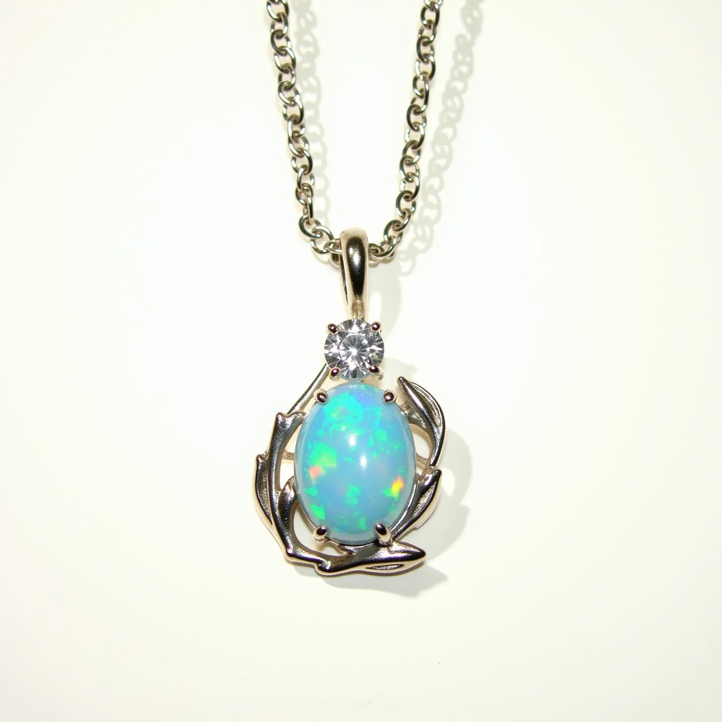 This opal necklace features a stunning, oval opal gemstone that showcases a vibrant play of colors, set in an ornate silver metal setting with a leaf motif that complements the centerpiece. Above the opal, a round-cut clear stone, likely a diamond or cubic zirconia, is set in a prong setting, adding a touch of sparkle to the design. The pendant is attached to a sleek chain that is likely made of silver, offering both elegance and durability. The necklace is secured with a simple yet secure clasp, ensuring it stays in place when worn. Its intricate design and combination of materials make it a unique and eye-catching piece.