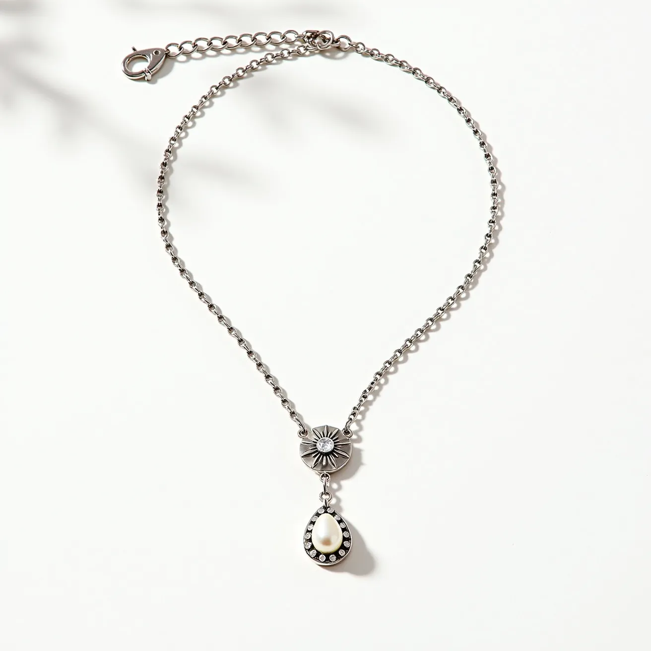 This pearl necklace features a distinct design with a pear-shaped pearl as its central element, elegantly set within a teardrop metal frame adorned with small round embellishments resembling gemstones. The necklace is complemented by a decorative connector featuring a floral or sunburst pattern, enhancing its artistic appeal. The chain is crafted from metal, characterized by small, evenly spaced links that contribute to its delicate look. A lobster clasp with an adjustable chain ensures a secure and customizable fit, making it both practical and stylish.