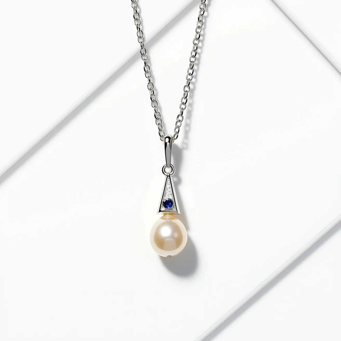 This pearl necklace features a solitary, lustrous pearl suspended below a triangular pendant. The pendant is adorned with a small, round blue gemstone, likely a sapphire, set within the triangle. The setting appears to be crafted from a polished metal, possibly silver or white gold, which complements the pearl's sheen. The chain is composed of oval links, securely attached to the pendant through a slender metal loop. The design is elegant, combining the timeless beauty of the pearl with the modern geometric accent of the triangular setting.