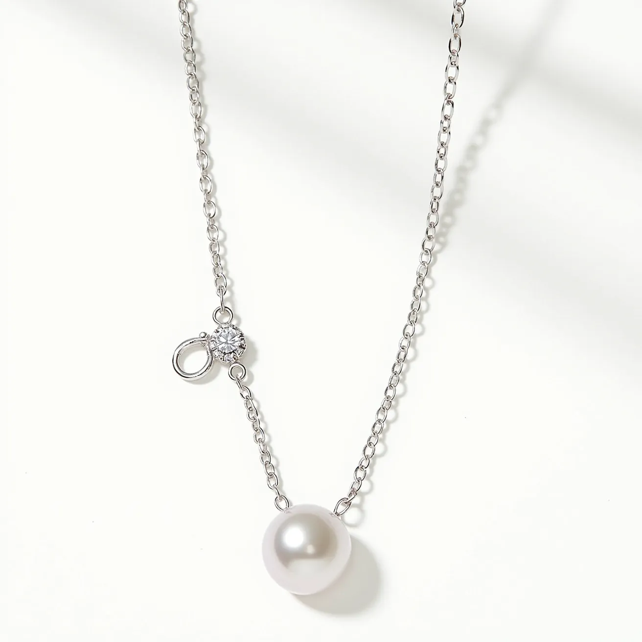 This pearl necklace features a single, lustrous white pearl as its centerpiece, elegantly suspended from a delicate silver chain. Alongside the pearl, there is a sparkling round clear gemstone, possibly a diamond, set in a classic prong setting. The necklace includes a simple yet secure clasp for fastening, contributing to its refined and graceful aesthetic. The materials and design of this necklace highlight its timeless beauty and sophistication.