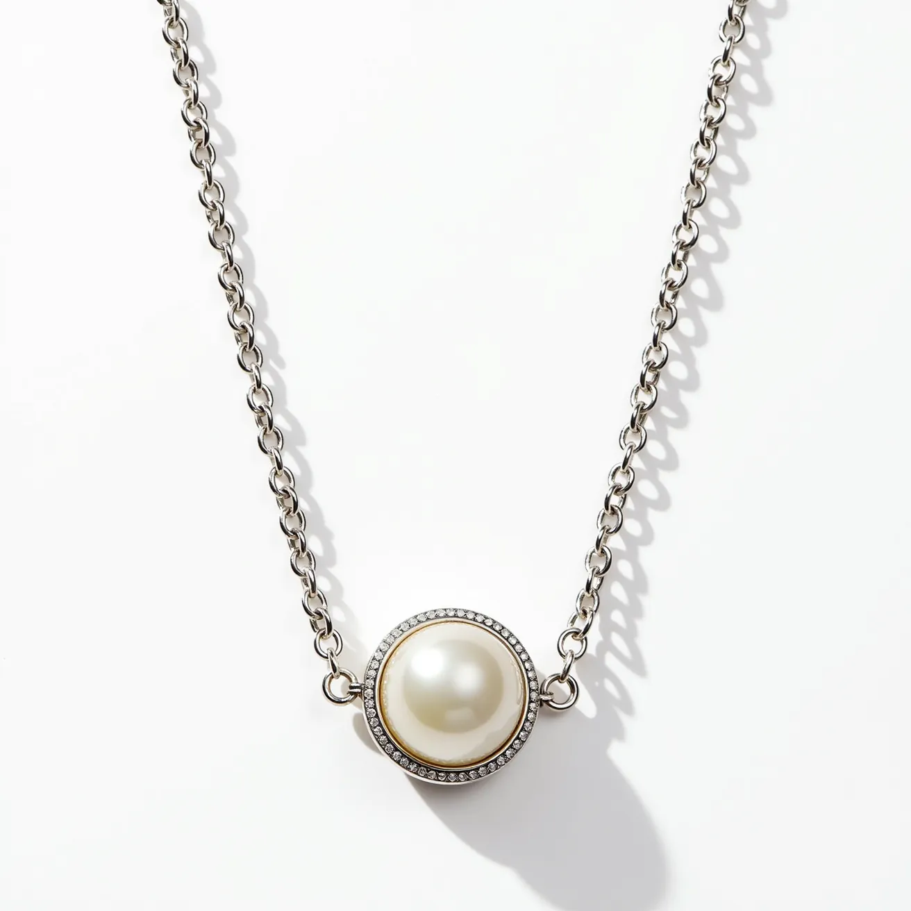 This pearl necklace showcases a single, large, luminous pearl set within a circular pendant. The pearl is elegantly encircled by a row of small, sparkling gems, likely diamonds, enhancing the pendant's brilliance and creating a stunning focal point. The pendant is attached to a metal chain with a polished finish, which provides a sleek and sophisticated look. The overall design is both classic and contemporary, highlighting the natural beauty of the pearl while adding a touch of glamour with the surrounding gems.