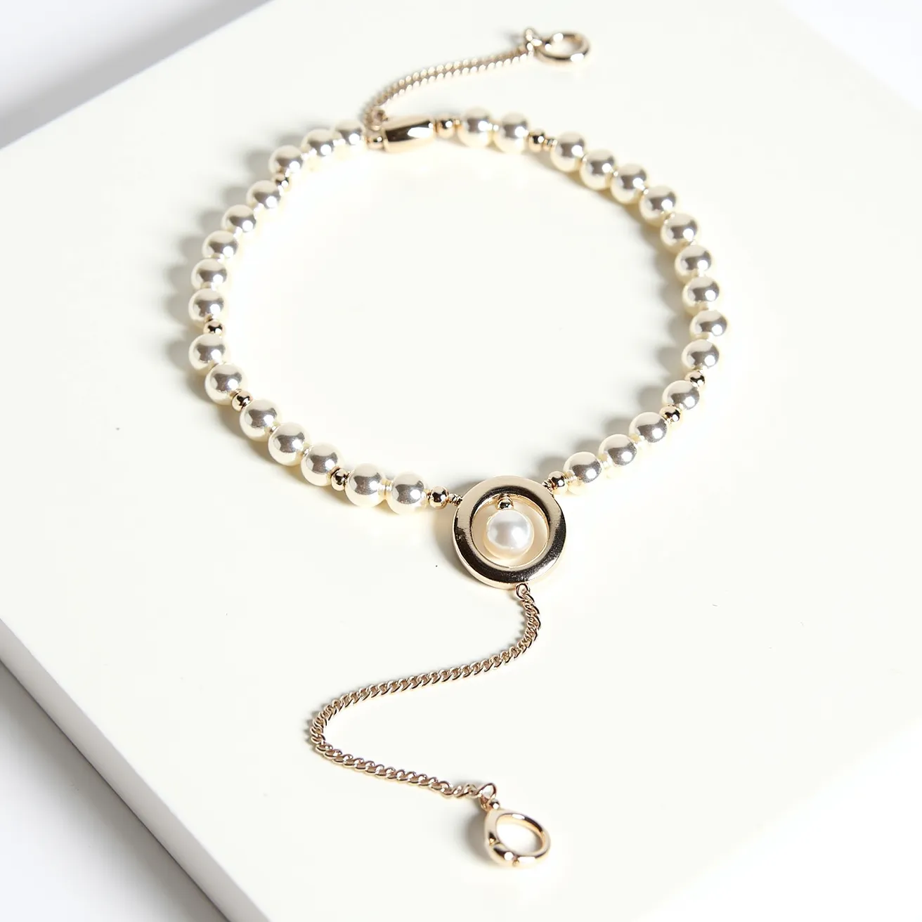 This pearl necklace features a series of lustrous, round pearls strung sequentially with small gold-tone spacer beads between each pearl. The central ornament showcases a single round pearl encased in a gold-tone circular setting, providing an elegant focal point. The necklace is secured by a lobster clasp, accompanied by an adjustable chain with a small loop at the end, facilitating versatile length adjustments. The combination of the classic pearls with modern gold-tone elements creates a timeless and sophisticated aesthetic.