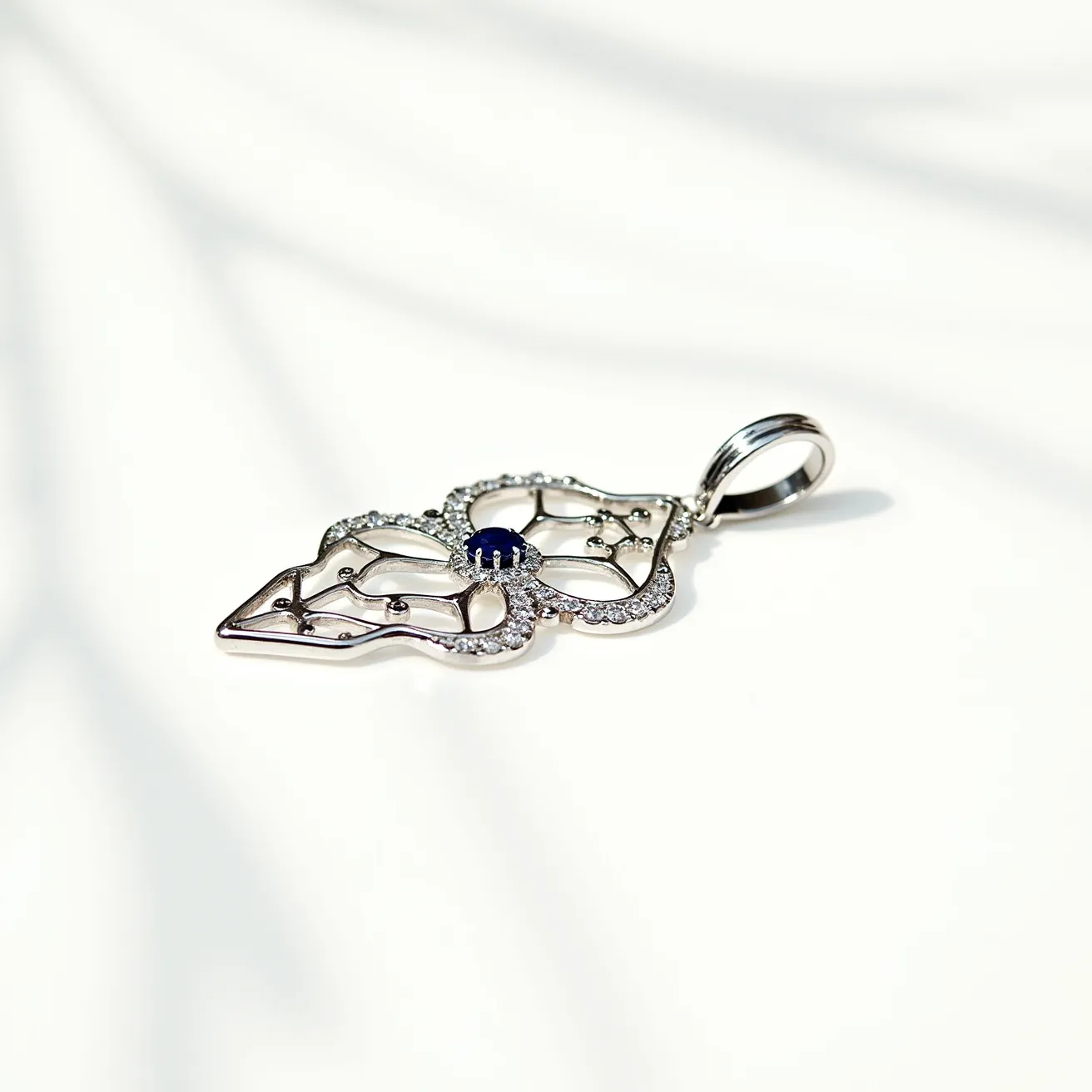 This statement pendant showcases an intricate openwork design crafted from a silvery metal, possibly platinum or white gold. A central, round-cut blue gemstone, likely a sapphire, is set in a secure prong setting at the heart of the pendant, surrounded by a halo of small, clear stones, potentially diamonds, set in a pavé style. The elegant shape of the pendant is emphasized by delicate curves and embellishments that enhance its sophisticated look. The pendant features a sturdy bail at the top, allowing it to be easily attached to a chain.