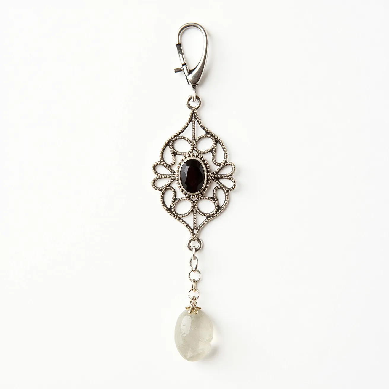 This statement pendant features a vintage-inspired design with an intricate metal framework, likely crafted from an antique-finished metal, giving it a classic aesthetic. At the center of the pendant is an oval-cut black gemstone, secured within a bezel setting, providing a striking focal point. The pendant also includes a dangling translucent teardrop-shaped stone, which adds an elegant touch to the piece. It is equipped with a metal clasp at the top, appearing robust and practical for attaching to various accessories. The overall design merges elegance with a hint of vintage charm.