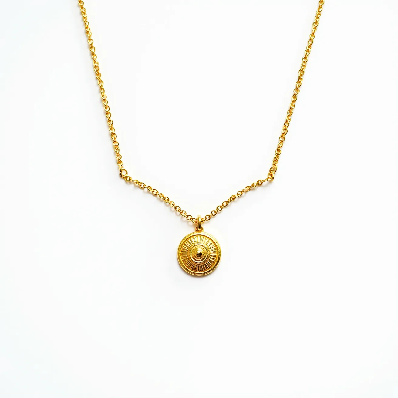 This pendant necklace features a delicate gold chain that elegantly supports a circular gold pendant. The pendant has intricate detailing radiating from the center, resembling sun rays, with a raised central design that adds depth and texture. Made from a polished gold material, the necklace reflects light beautifully and enhances its luxurious appearance. The chain is linked skillfully, suggesting durability and style, and is secured with a small loop at the pendant's top. The clasp is not visible here, implying a continuous elegance that promises comfort and ease of wear.