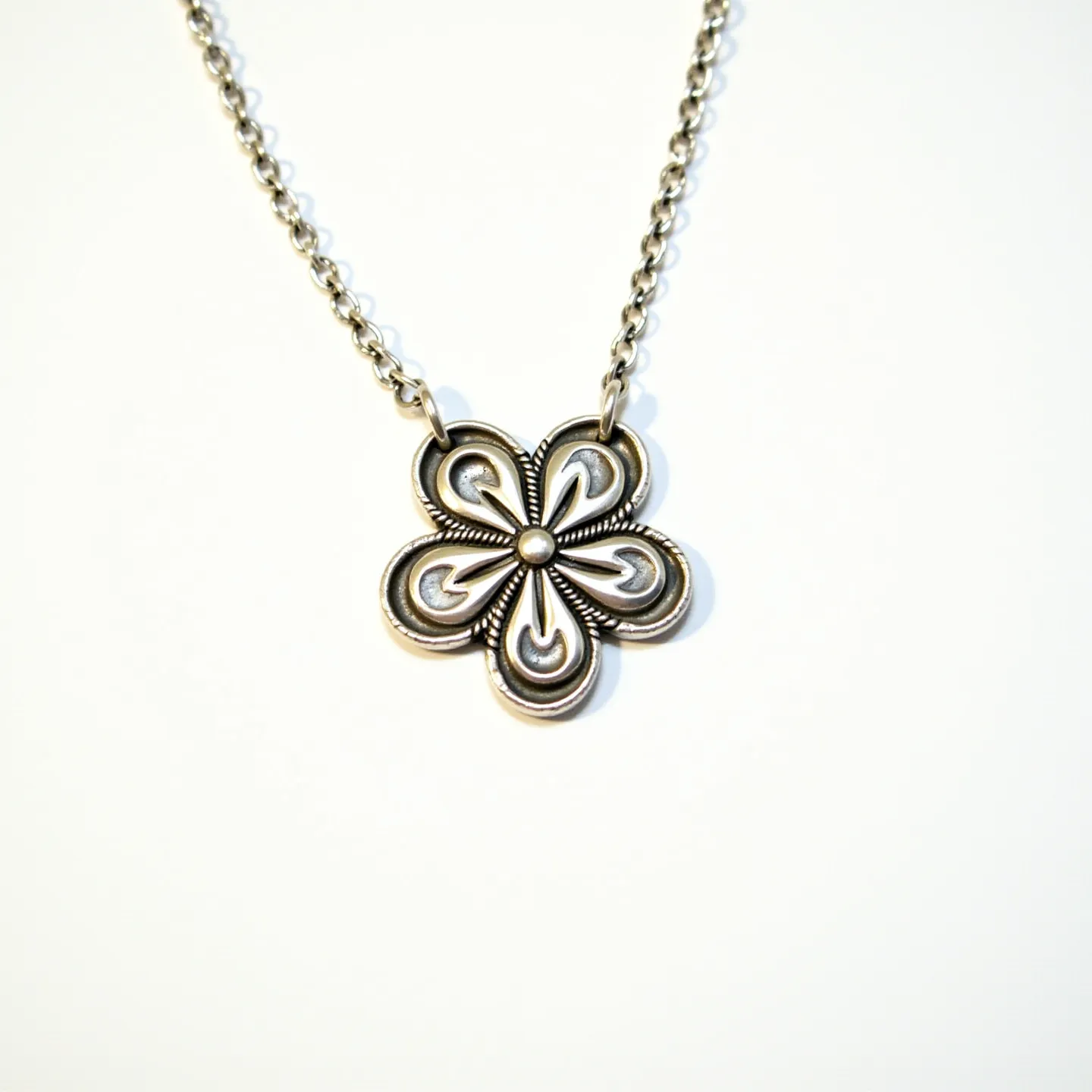 This pendant necklace features a delicate floral design, crafted from a silver-toned metal that gives it a vintage aesthetic. The flower shape, intricate with loops and curves, is highlighted by a central raised dot detail. The pendant is suspended from a matching chain, which appears to be made of small, closely linked metal loops, adding to the cohesive look of the piece. The chain likely secures with a standard clasp, ensuring ease of wear and security. The pendant's artistic design and the subtle shine of the metal come together to create a timeless accessory.