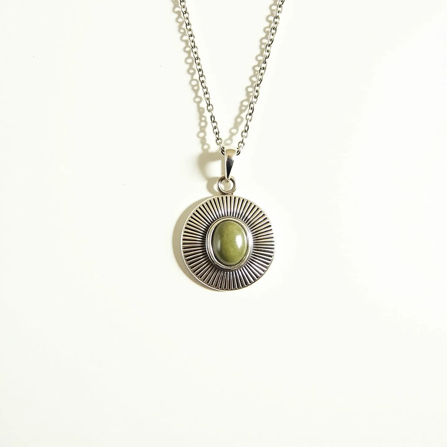 This pendant necklace features a central olive-green oval stone, possibly jade, set in a bezel setting. The stone is surrounded by a circular silver-toned metal base with radiating etched lines, creating a sunburst effect. The pendant is attached to a delicate, silver-toned chain with interlocking round links. The chain is fastened with a traditional spring ring clasp, ensuring a secure fit. This piece showcases a combination of earthy and metallic elements, offering a stylish and sophisticated look.