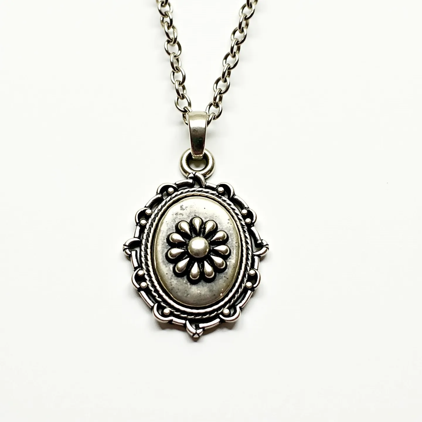 This pendant necklace features an intricately designed oval-shaped pendant made of metal, likely silver or a silver-toned alloy. The centerpiece of the pendant is a raised floral motif with a central bead and petal-like embellishments, surrounded by a decorative border that enhances its vintage aesthetic. The pendant is suspended from a sturdy chain with interlocking links, suggesting durability and classic style. The attachment to the chain is facilitated by a simple bail, allowing the pendant to hang gracefully. The necklace is secured with a standard clasp, which complements its overall elegant design.