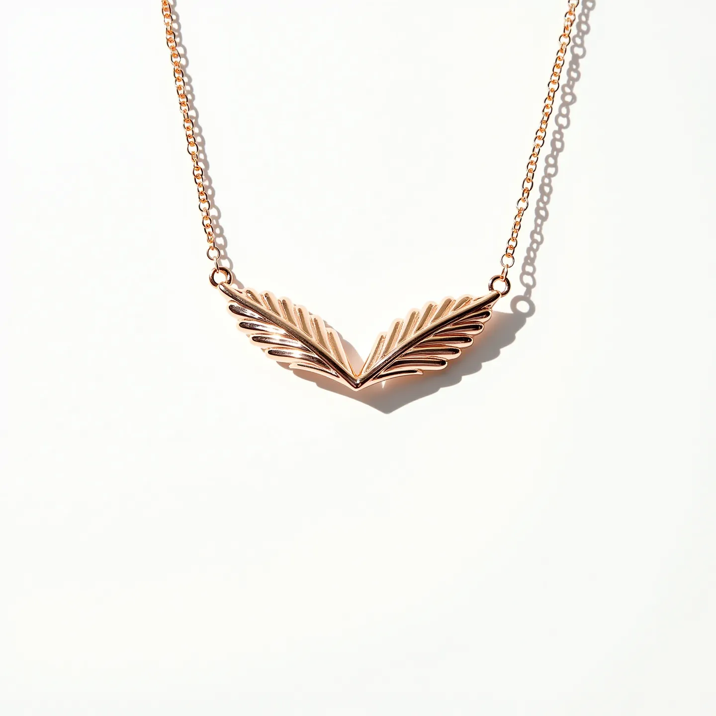 This pendant necklace features a striking design that showcases a pair of symmetrical feather-like elements crafted from what appears to be a polished rose gold metal. The feathers converge centrally, forming a gentle V-shape that highlights the symmetry and elegance of the piece. Each feather exhibits a detailed texture, enhancing the natural motif and adding dimension to the pendant. The necklace is suspended from a delicate chain, also in a rose gold hue, that complements the pendant and is likely secured with a simple clasp, providing both functionality and style.