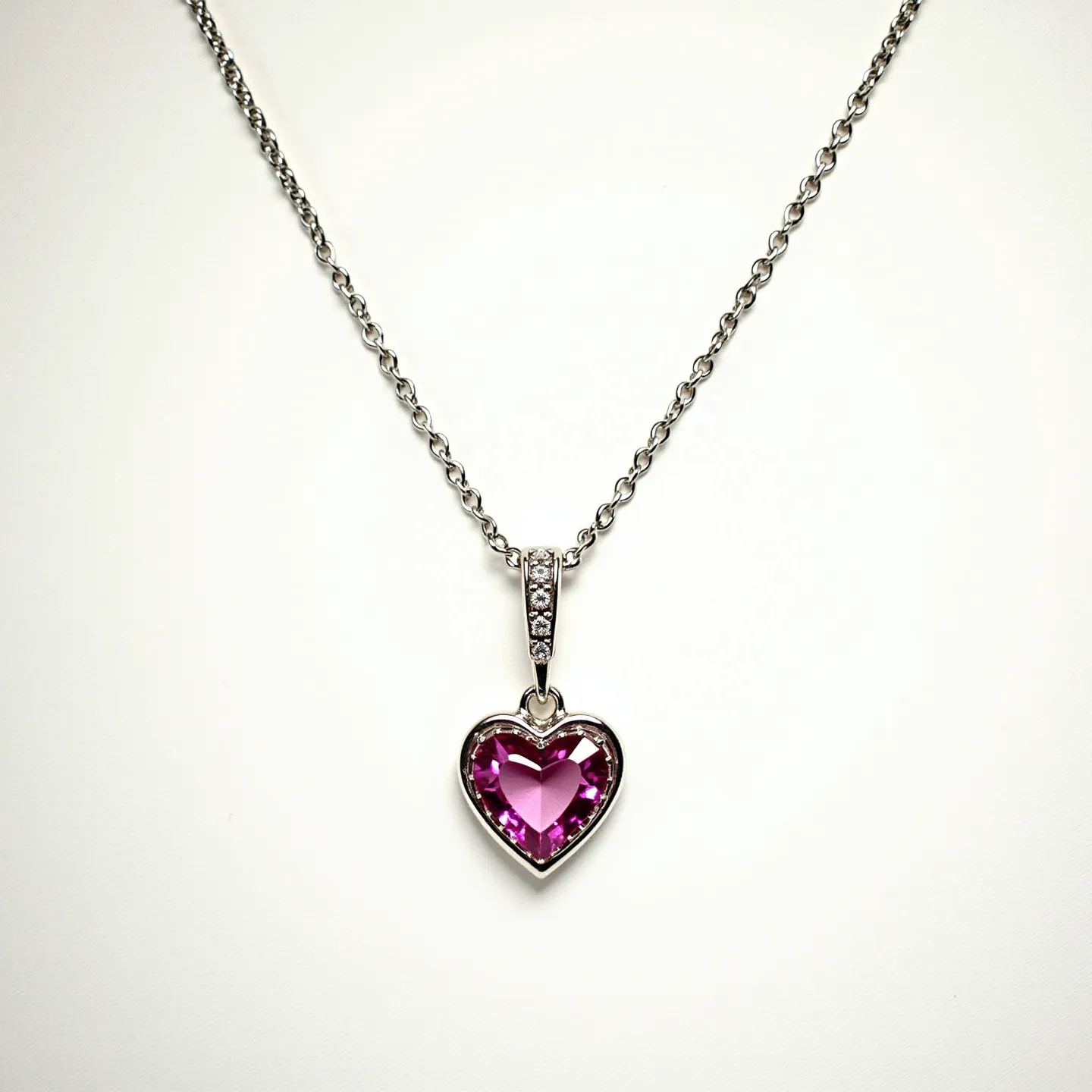 This pink heart necklace features a delicate silver chain paired with a striking heart-shaped pink gemstone. The gemstone is likely a pink sapphire or topaz, cut into a heart shape to maximize its sparkle and visual appeal. It is encased in a simple but elegant silver setting that accentuates the stone's vibrant color. Above the gemstone, the pendant is attached to the chain through a decorative bail adorned with small, clear stones, possibly diamonds or cubic zirconia, adding a touch of brilliance. The chain appears to have a standard clasp, ensuring secure wearability.