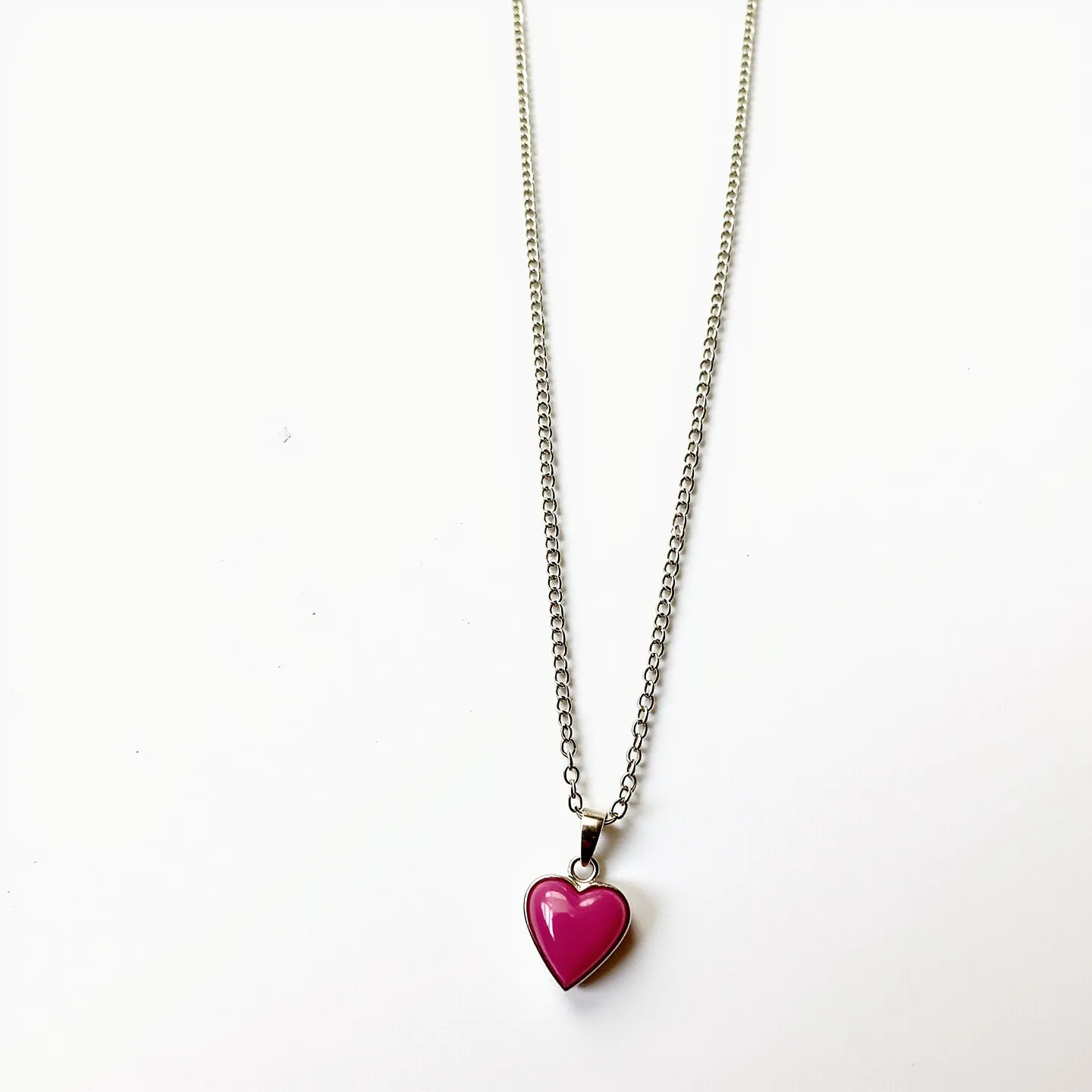 This pink heart necklace features a delicate chain, likely made of a metal such as sterling silver or stainless steel, with a polished finish. The pendant is a heart-shaped piece composed of a bright pink material, possibly enamel or a synthetic stone, with a smooth, glossy surface. It is set in a simple bezel setting that complements its vibrant color. The necklace connects with a subtle clasp, typically a lobster clasp or spring ring, which secures the piece comfortably around the neck. The chain's uniform links add a classic touch to the overall design.