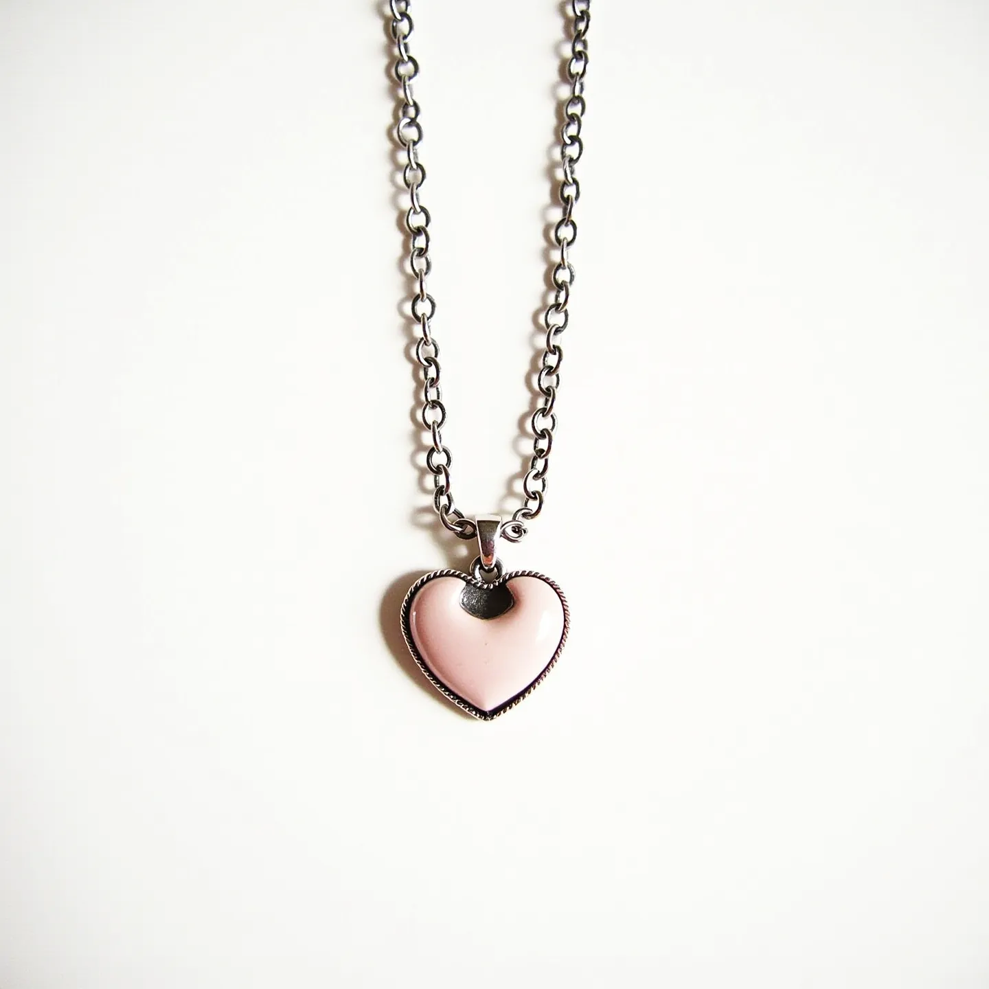 This pink heart necklace features a heart-shaped pendant in a soft pink hue, likely made from enamel or a pink gemstone. The pendant is encased in a sleek metal setting, possibly silver or stainless steel, with a decorative rope-like edge that enhances its visual appeal. The pendant hangs from a sturdy chain comprised of oval links, suggesting durability and a classic style. The attachment of the pendant to the chain is facilitated by a small, smooth bail, ensuring secure connectivity while allowing gentle movement. The necklace closes with a standard clasp, likely a lobster clasp, providing a reliable fastening mechanism.