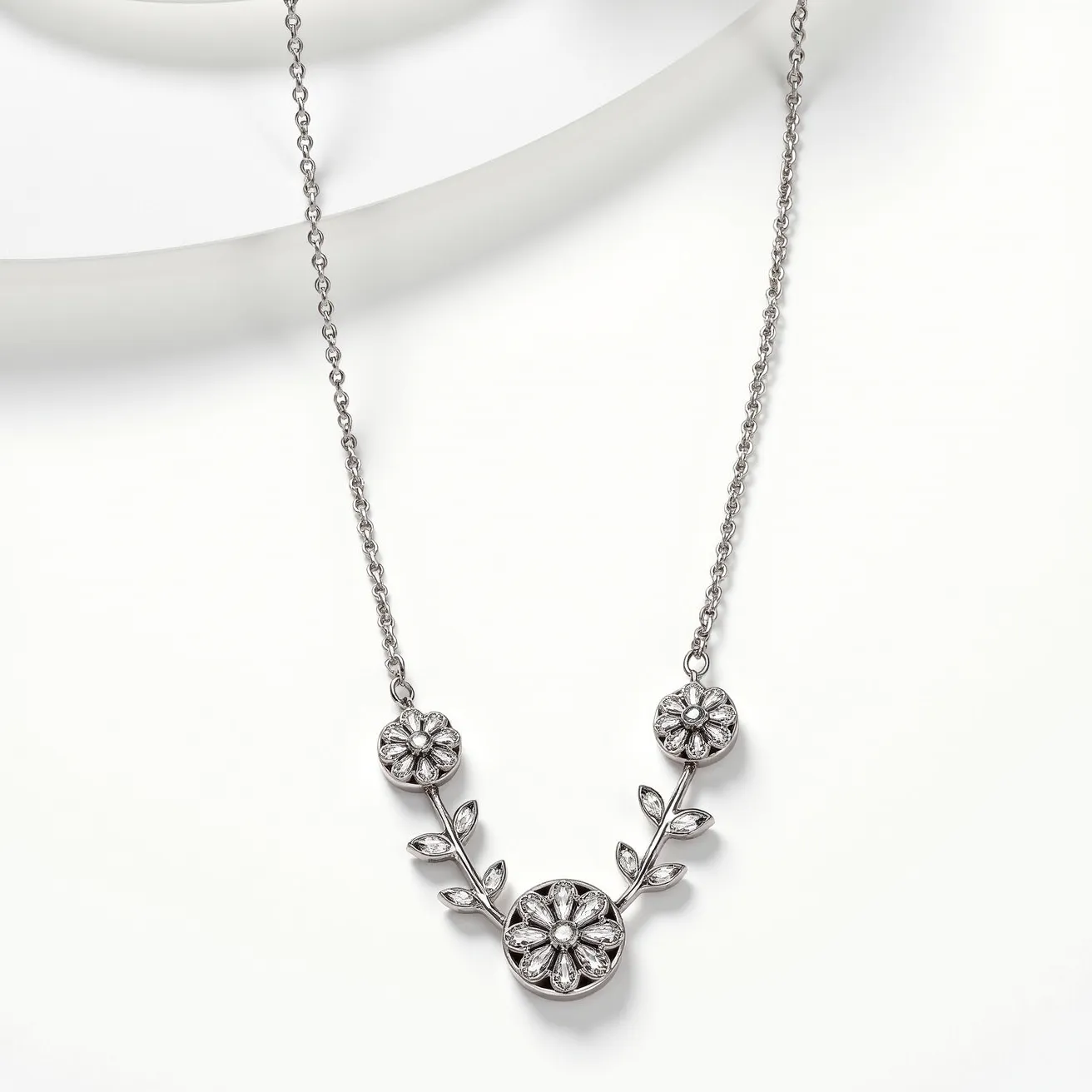 This platinum necklace features a delicate, elegant design with intricate floral and leaf motifs. At the center are three flower-shaped elements, each adorned with sparkling gems set in a prong-like style that highlights their brilliance. The central flower is slightly larger, serving as the focal point, while the smaller ones complement it on either side. Each flower is accentuated with diamond-like gems that have a round and marquise cut, creating a shimmering effect. The flowers are connected by a branch-like structure with leaf motifs, enhancing the naturalistic theme. The chain is finely crafted, adding to the overall sophistication of the piece, and the necklace likely fastens at the back with a secure clasp, ensuring both style and functionality.