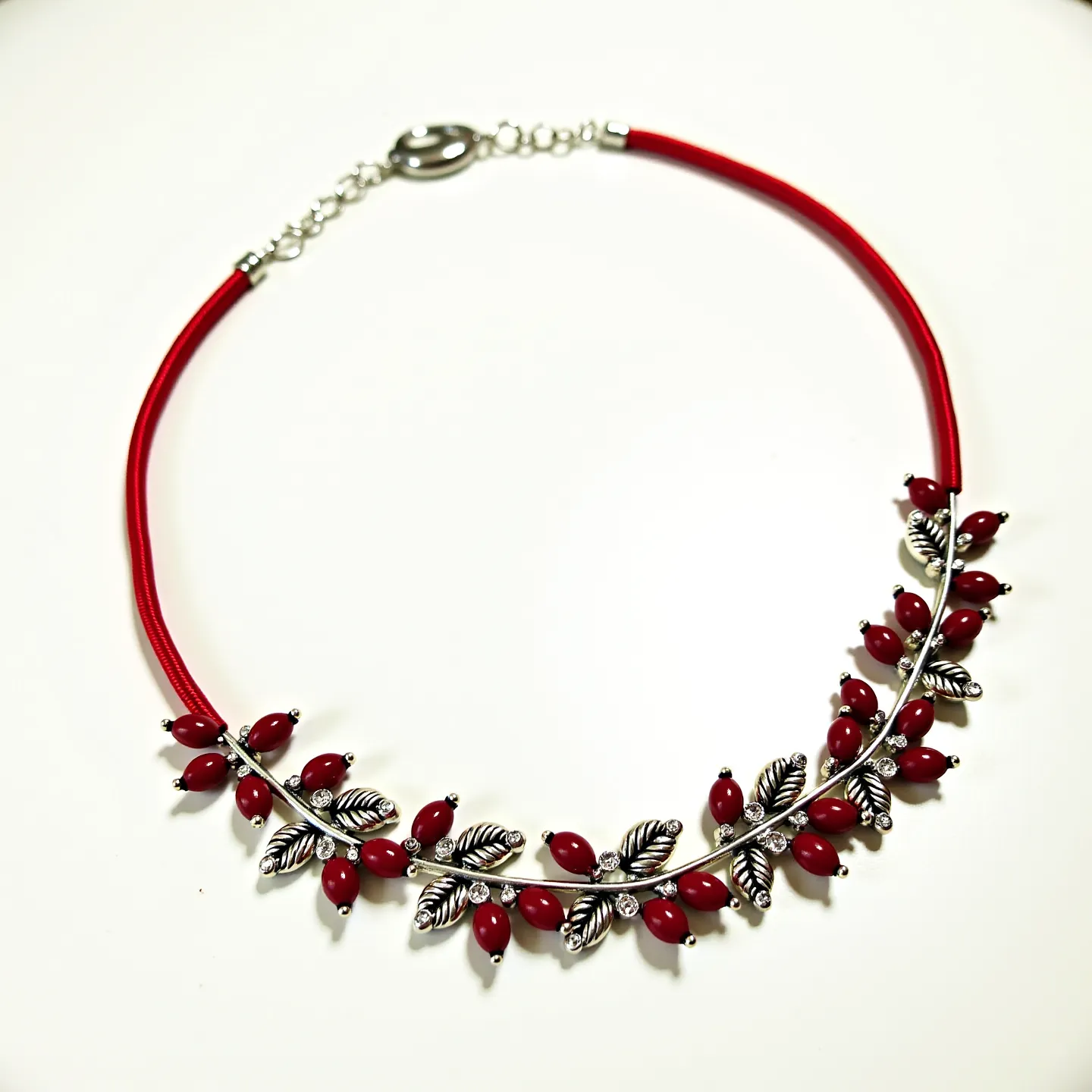 This red necklace features an elegant design with red, oval-shaped bead-like stones paired with silver-toned leaf accents. The red stones are uniformly distributed along a metallic arc, each held in place with a simple prong setting. Small, clear round stones add a subtle sparkle between the leaves and red beads. The necklace is finished with a lobster clasp and a short chain, allowing for adjustable length. The bright red cord adds a bold contrast to the metallic embellishments, creating an appealing blend of texture and color.