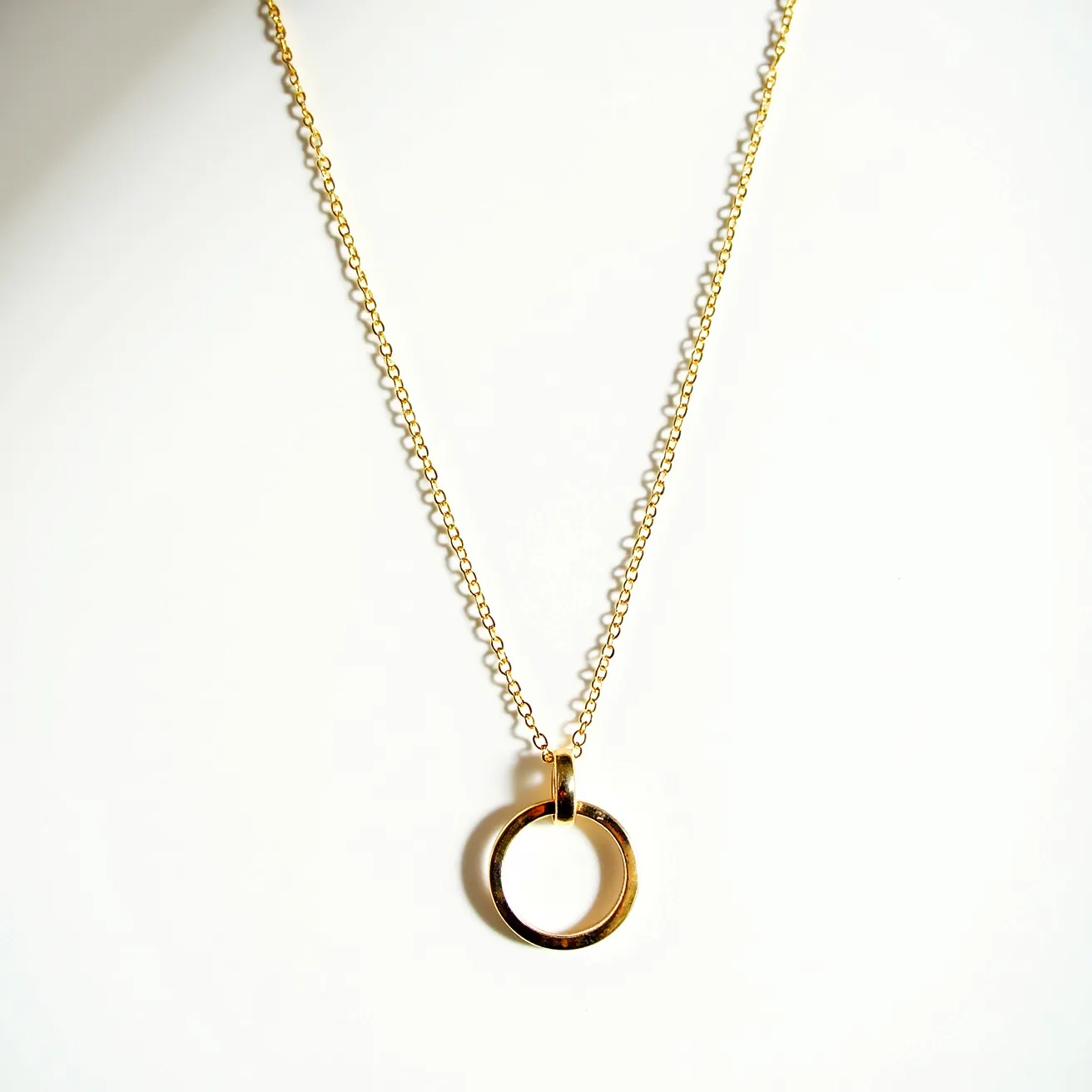 This ring necklace features a delicate gold chain and a pendant in the shape of a simple, polished gold ring. The pendant is attached to the chain with a small gold loop, seamlessly integrating it into the necklace. The chain consists of small, interlocking links that provide a refined and elegant appearance. The necklace likely uses a clasp mechanism common in jewelry, though the specifics of the clasp are not visible in the image. Overall, the design reflects a minimalist aesthetic, emphasizing the beauty of the gold material.