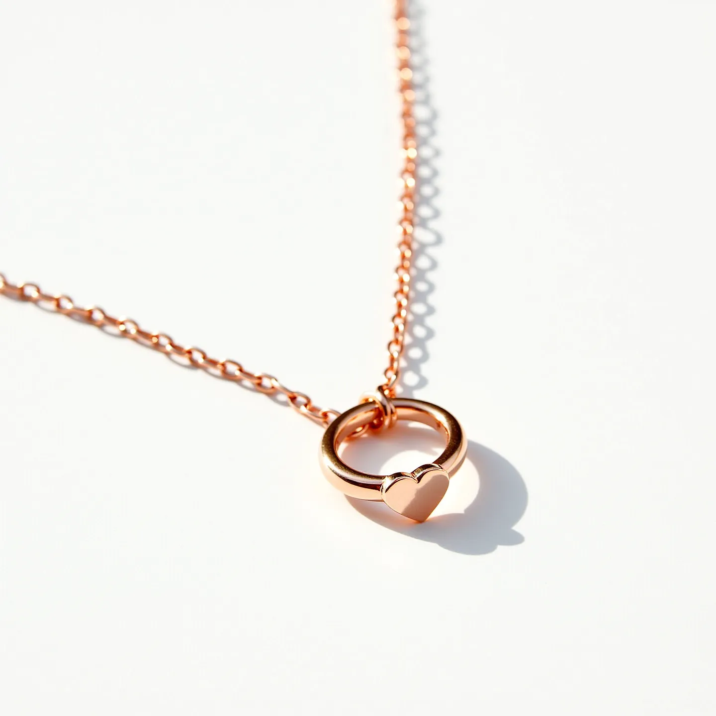 This ring necklace features a polished rose gold finish, showcasing a circular pendant with a smooth heart embellishment on its lower section. The chain is composed of evenly linked rose gold elements, offering a consistent and elegant appearance. The pendant hangs freely, connected securely through a simple loop, ensuring fluid movement along the chain. There are no visible gemstones or intricate stone settings present in the design. The necklace likely utilizes a typical clasp mechanism, such as a lobster or spring ring, to fasten comfortably around the neck.