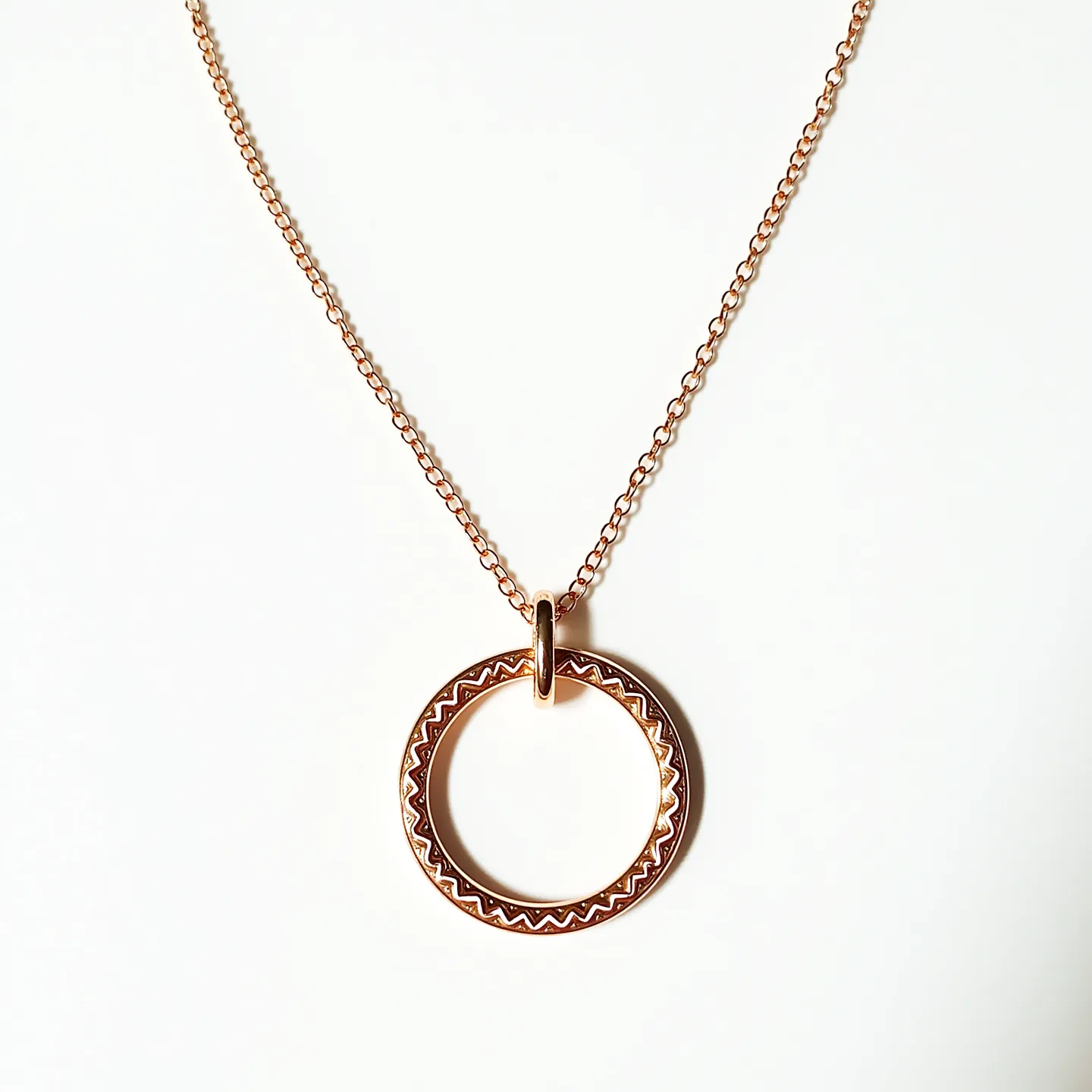 This ring necklace features a circular pendant crafted from what appears to be gold or gold-toned metal, displaying intricate zigzag engravings along its surface, adding an element of texture and design. The pendant is suspended from a matching gold or gold-tone chain, likely composed of fine cable links, providing a delicate and elegant look. The chain is attached to the pendant through a smooth, polished loop that complements the overall aesthetic. There are no visible gems or stones, and the necklace likely employs a simple spring-ring or lobster clasp to facilitate wearing.