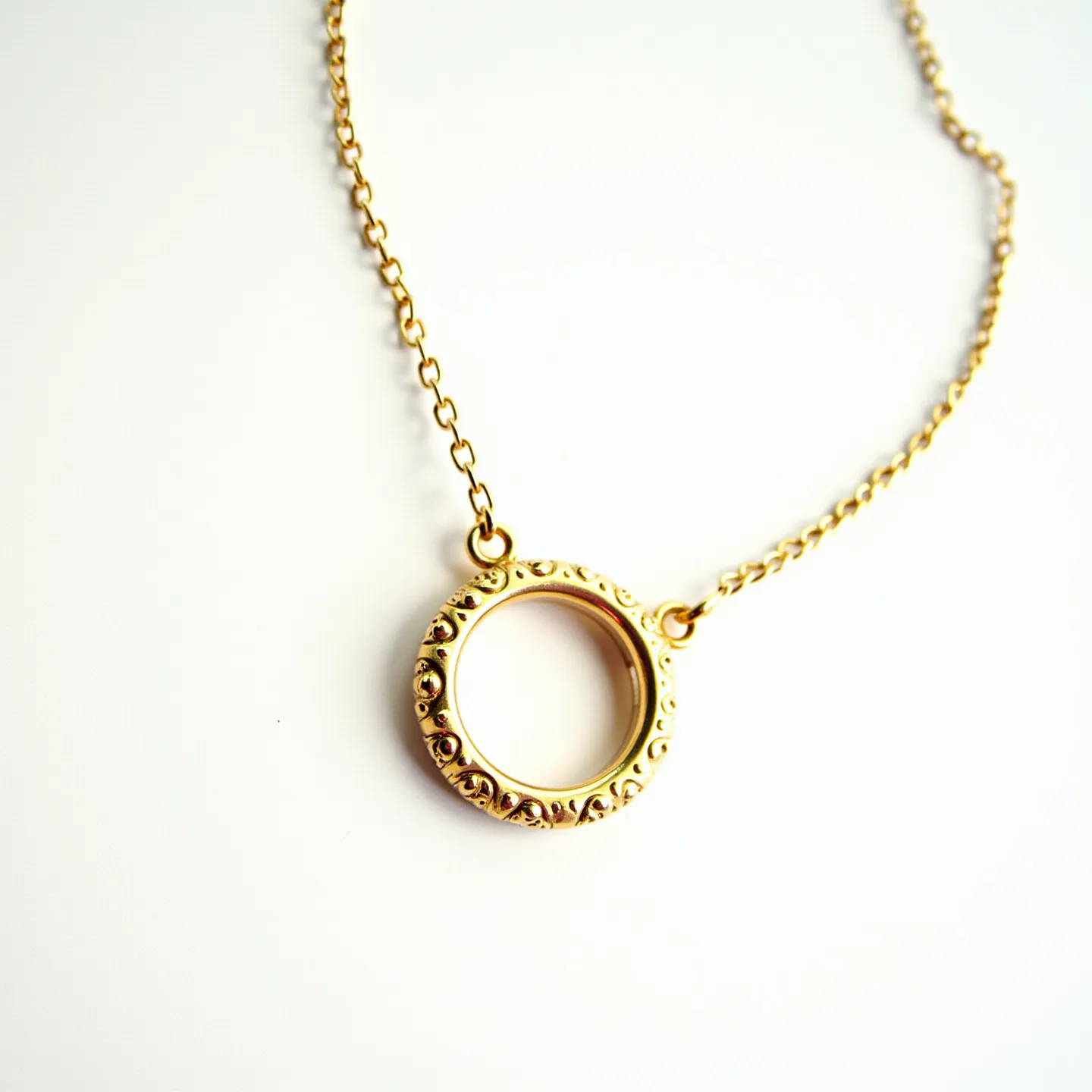 This ring necklace features a circular pendant crafted from a gold-toned metal, showcasing intricate detailing around its perimeter that adds texture and visual interest. The pendant is seamlessly integrated into a matching delicate chain, which is composed of evenly spaced links, contributing to an elegant and cohesive design. The pendant’s surface is smooth and lustrous, with no embedded gems or stones, focusing the attention on its ornate craftsmanship. The necklace is designed with a simple yet effective attachment mechanism, ensuring secure wear while accentuating the minimalist aesthetic of this piece.