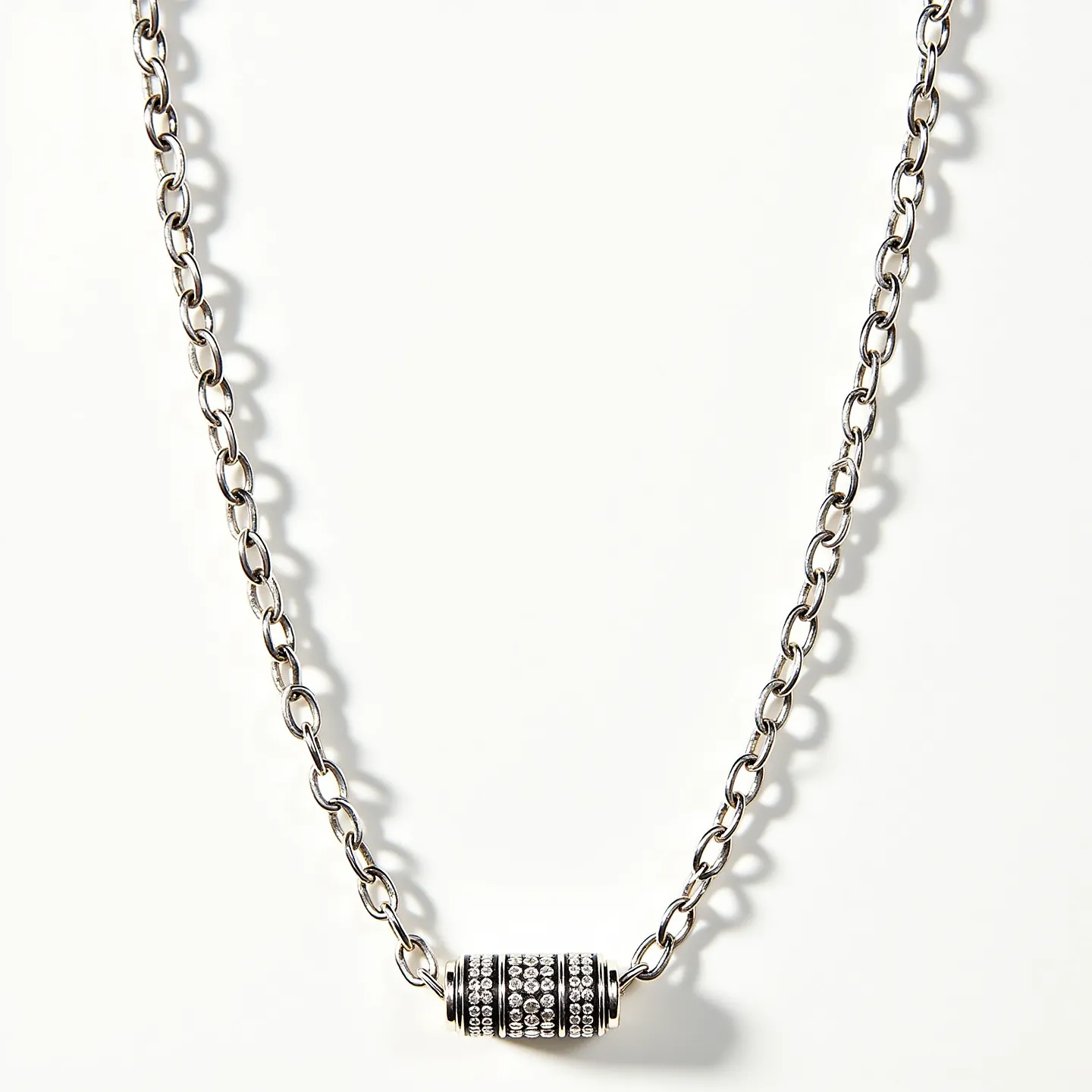 This rolo chain necklace features a series of interconnected round links, likely made from a polished metal such as silver or stainless steel, giving it a sleek and modern appearance. The necklace is adorned with a cylindrical pendant that is encrusted with numerous small, shimmering gemstones, possibly resembling diamonds or crystals, set in rows to enhance its sparkle. The pendant is designed to catch light, adding a touch of elegance and sophistication to the piece. The necklace appears to include a standard clasp, ensuring secure wearability while complementing its overall aesthetic.