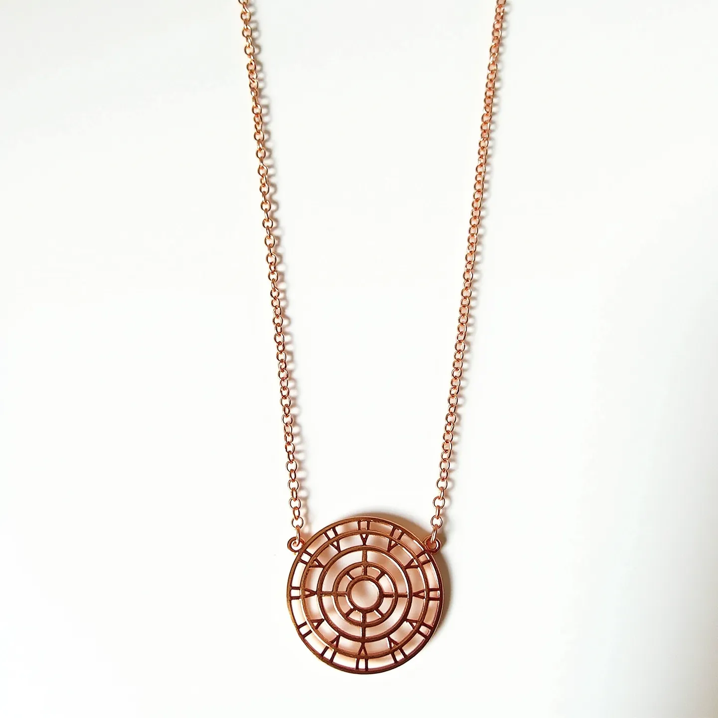 This rose gold necklace features a delicate chain which holds a circular pendant with an intricate grid pattern design. The necklace is crafted from rose gold, giving it a warm and elegant hue. The pendant is composed solely of rose gold without any gemstones, emphasizing its minimalist geometric design. The chain is a simple link style, contributing to the necklace's sleek appearance. The piece likely includes a standard clasp mechanism at the back, providing a secure fastening for everyday wear.