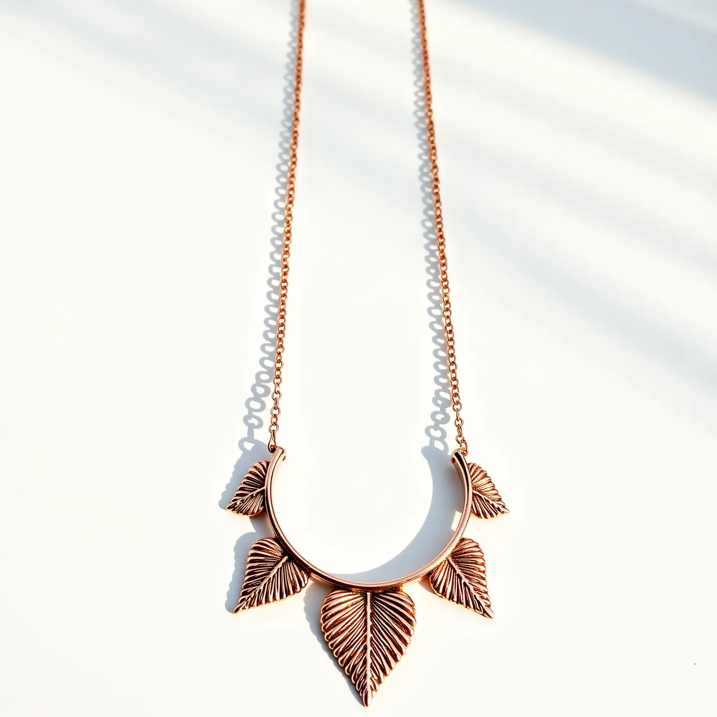 This rose gold necklace features an elegant design with intricately detailed leaf-like charms hanging from a polished, crescent-shaped bar. The chain itself is composed of delicate links that complement the warm hue of the rose gold. The leaf charms are artistically crafted with fine lines that mimic the natural veins of a leaf, adding texture and visual interest to the piece. The necklace is secured with a standard lobster clasp, ensuring ease of wear and security. The overall design combines natural motifs with a modern aesthetic, making it both sophisticated and versatile.